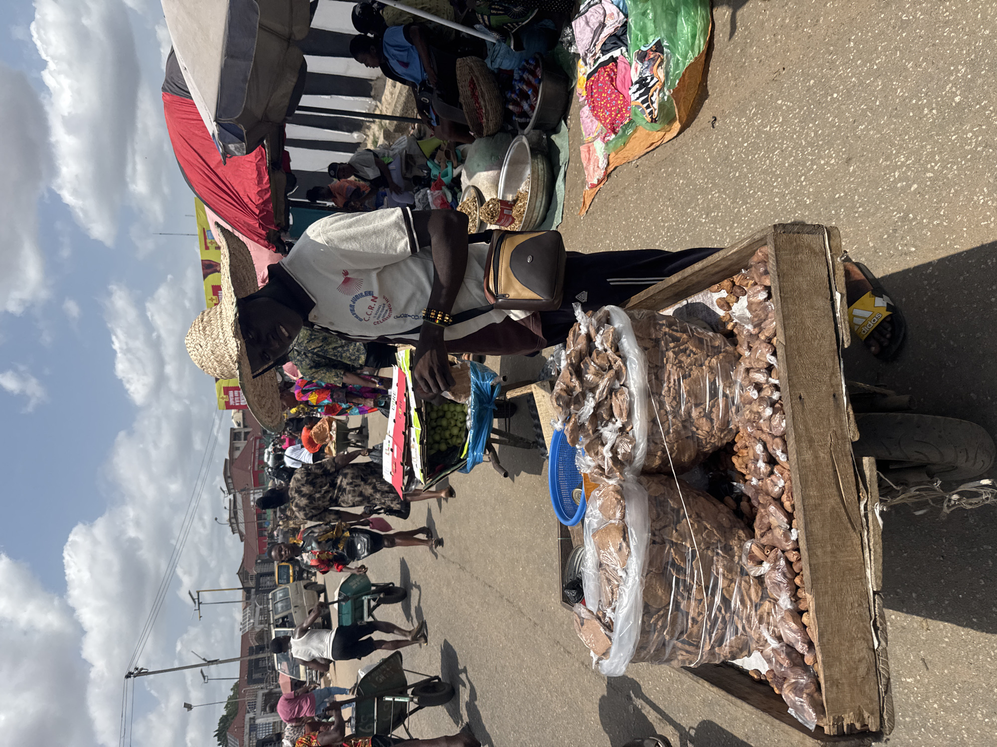 This media file depicts a vibrant and bustling street market scene in Onitsha, Nigeria, under bright daylight with a clear sky dotted with white clouds.
The central figure is a male vendor, appearing to be in his 30s or 40s, wearing a straw hat, a white t-shirt with a red phoenix-like logo and text reading "JUBILEE C.C.R.N CELEBRATI...", dark pants, and a brown shoulder bag. He has a beaded bracelet on his left wrist. He is standing next to a wooden pushcart heavily loaded with numerous clear plastic bags, each filled with light brown, irregularly shaped items, likely nuts, dried fruits, or local snacks. A blue plastic basket also sits on the cart.
Behind him and extending down the street, the market is alive with activity. Other vendors are present, some under colorful awnings (red and grey), displaying various goods including textiles and produce. A blue bag filled with green, spherical fruits (possibly apples or similar) is visible near the main vendor's cart. Numerous people, including other vendors and shoppers, can be seen walking, standing, and interacting. Several pushcarts and vehicles, such as vans and motorcycles, are also on the paved street. Buildings with distinct architecture and utility poles line the background, indicating an urban setting. A yellow sandal with the "adidas" logo is visible on the ground near the cart. The scene conveys a sense of everyday commerce and community in a busy market environment.