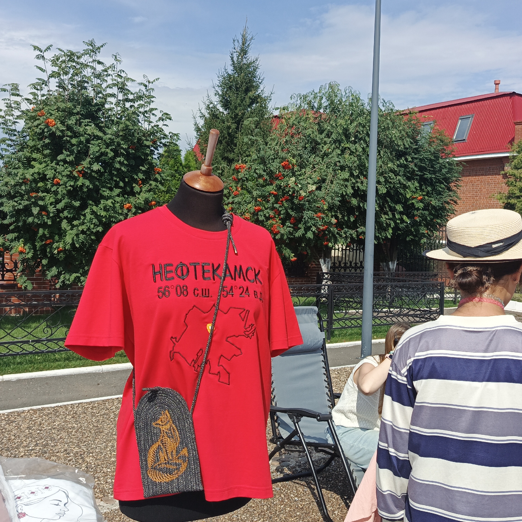 The image depicts an outdoor market scene in Nikolo-Beryozovka, Russia. In the foreground, a red t-shirt is displayed on a mannequin. The t-shirt features black text that reads "НЕФТЕКАМСК" (Neftekamsk) and coordinates "56°08 с.ш. 54°24 в.д." (56°08 N.L. 54°24 E.L.), along with a map outline. A small, grey woven bag with an embroidered orange fox design is hanging from the mannequin's neck. To the right, a person wearing a straw hat with a black ribbon is seen from behind, facing away from the camera. They are wearing a striped blue and white shirt. Another person, partially visible, is seated in a grey folding chair in the background. The environment includes trees with red berries, a red-roofed brick building, a black metal fence, and a grey pole. The ground is covered with gravel. The sky is blue with some clouds.