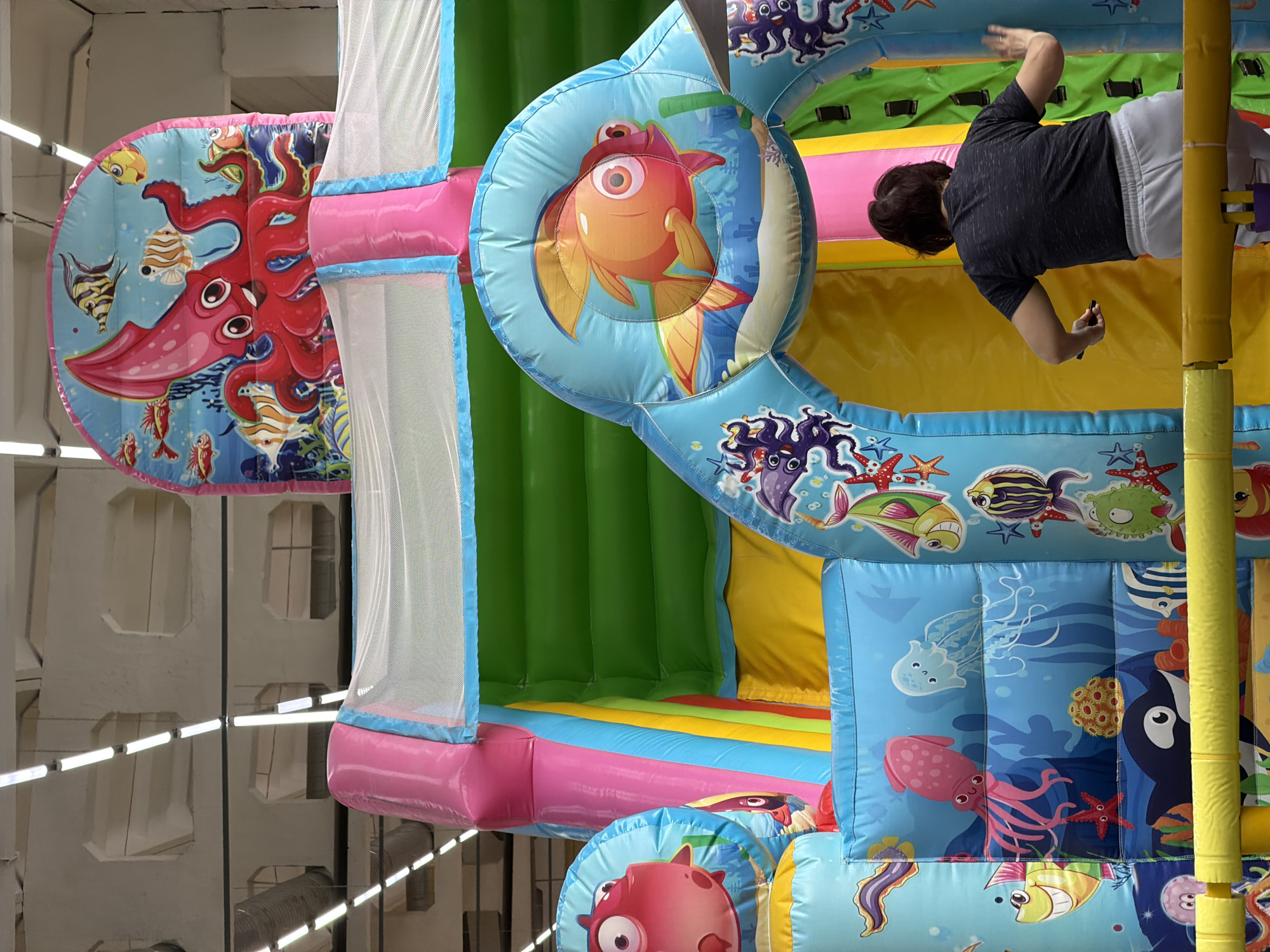 A child, wearing a gray shirt and white shorts, is inside a multi-colored inflatable play structure. The structure is decorated with images of sea creatures, including octopuses, fish, and jellyfish. The play structure is located indoors, possibly in a play area or entertainment center. The location is Chaikovsky, Russia. The environment includes windows and ceiling lights, suggesting an indoor setting. The child appears to be interacting with the structure, possibly playing or climbing.