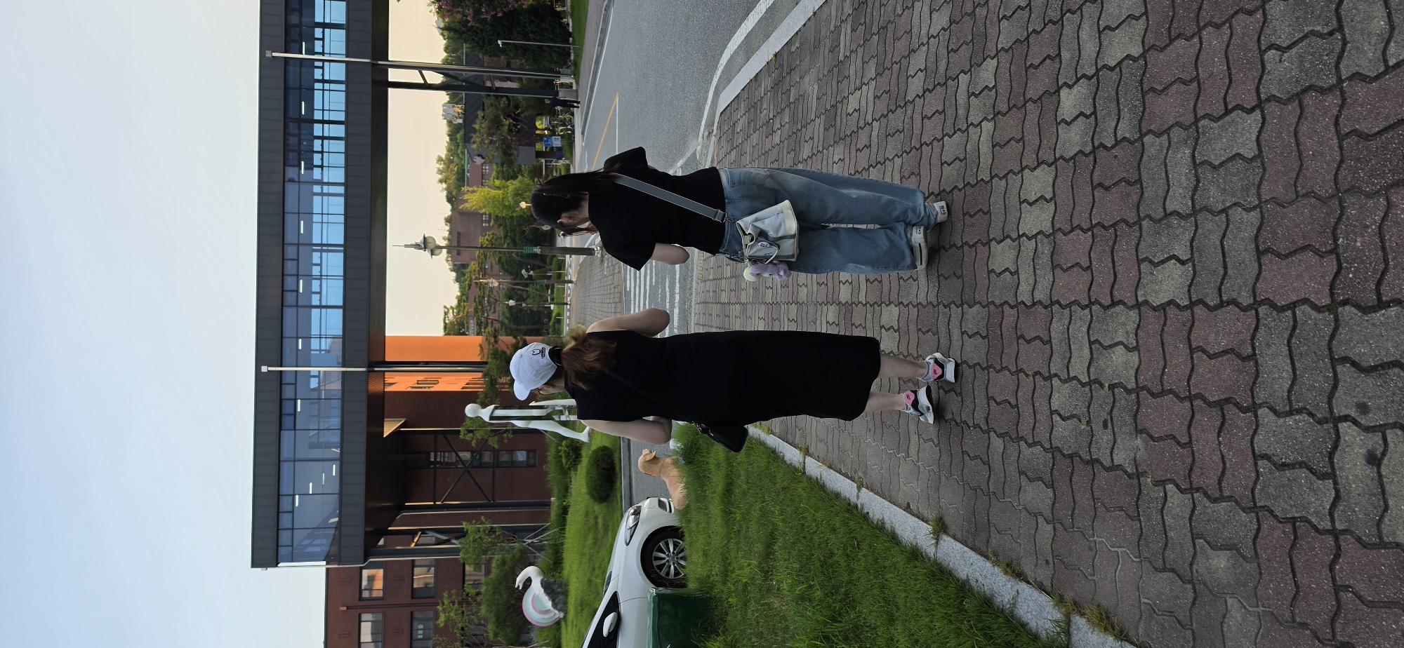 Two women are walking away from the camera on a brick sidewalk. The woman on the left is wearing a black sleeveless dress and a white baseball cap. The woman on the right is wearing a black t-shirt and light blue jeans. Both women are wearing sneakers. A light-colored car is parked to the left of the sidewalk, with a light brown dog lying on the grass in front of it. In the background, there is a modern building with a sky bridge connecting two sections. The environment appears to be an outdoor campus or park area with trees and streetlights. The sky is clear and overcast.