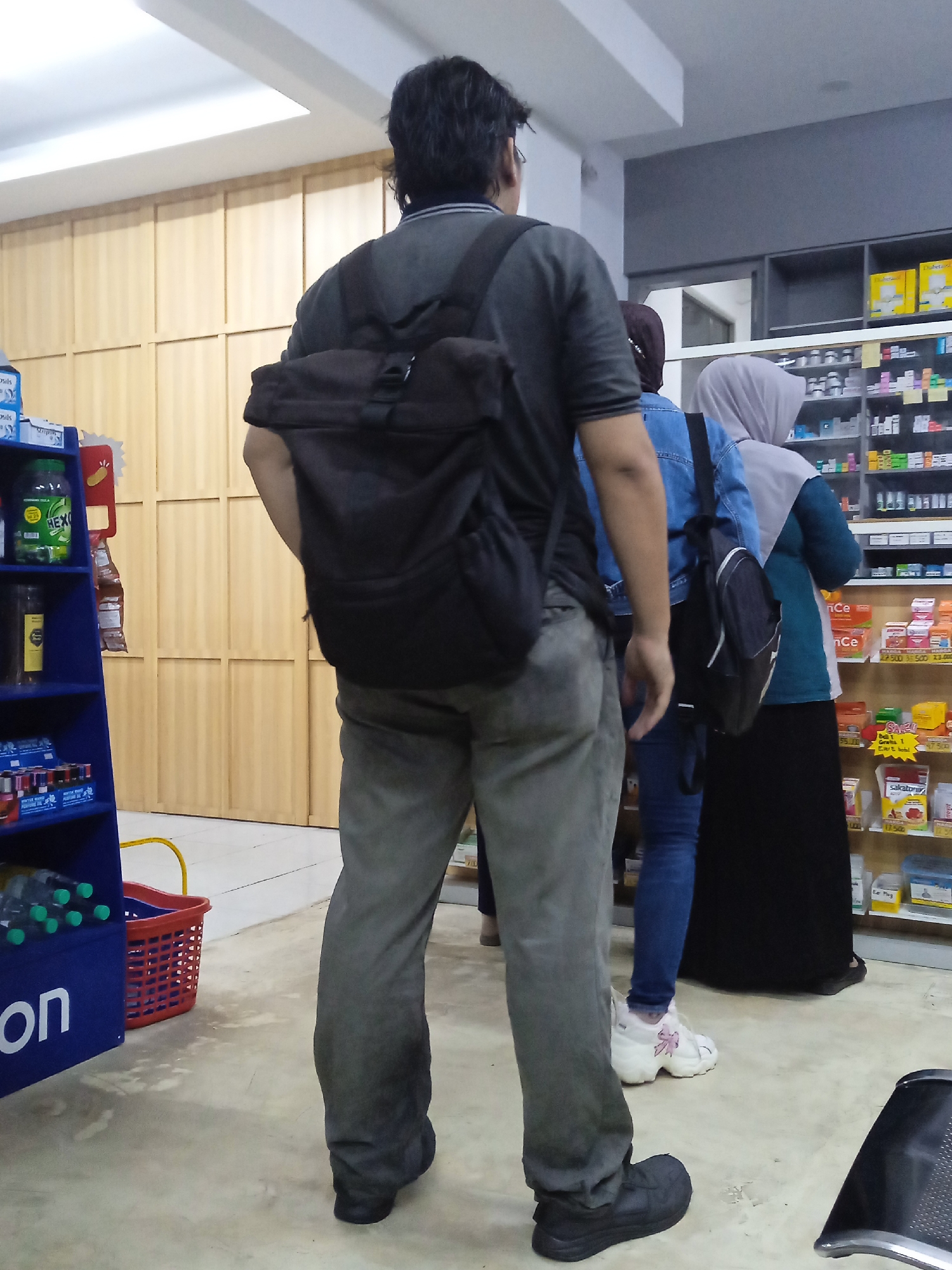 The image displays an indoor scene, likely a pharmacy or retail store in Kebonsari, Indonesia, featuring three individuals seen from behind. The person in the foreground, facing away from the camera, wears a dark grey short-sleeved shirt, grey pants, and black shoes, with a black backpack. To their right, a second individual is partially visible, wearing a blue denim jacket, blue jeans, and white sneakers with a pink bow, also carrying a black backpack. Further right, a third person is seen wearing a light grey hijab, a teal top, and a black skirt.

The setting includes extensive shelving stocked with various products. To the left, a blue display unit holds items, including green bottles labeled "HEXI" and boxes with "sils" printed on them. A red plastic shopping basket rests on the light-colored floor. Behind this display is a light wooden paneled wall. To the right, numerous white shelves are densely packed with medications, health products, and other goods. Several items feature price tags with values such as "27.500," "38.500," "23.000," and "17.500." A red "SALE!" tag is also visible. Text on product packaging includes "Ce," "nCe," "sakatori," and "Dabet." The scene suggests customers waiting or browsing in a well-lit store environment.