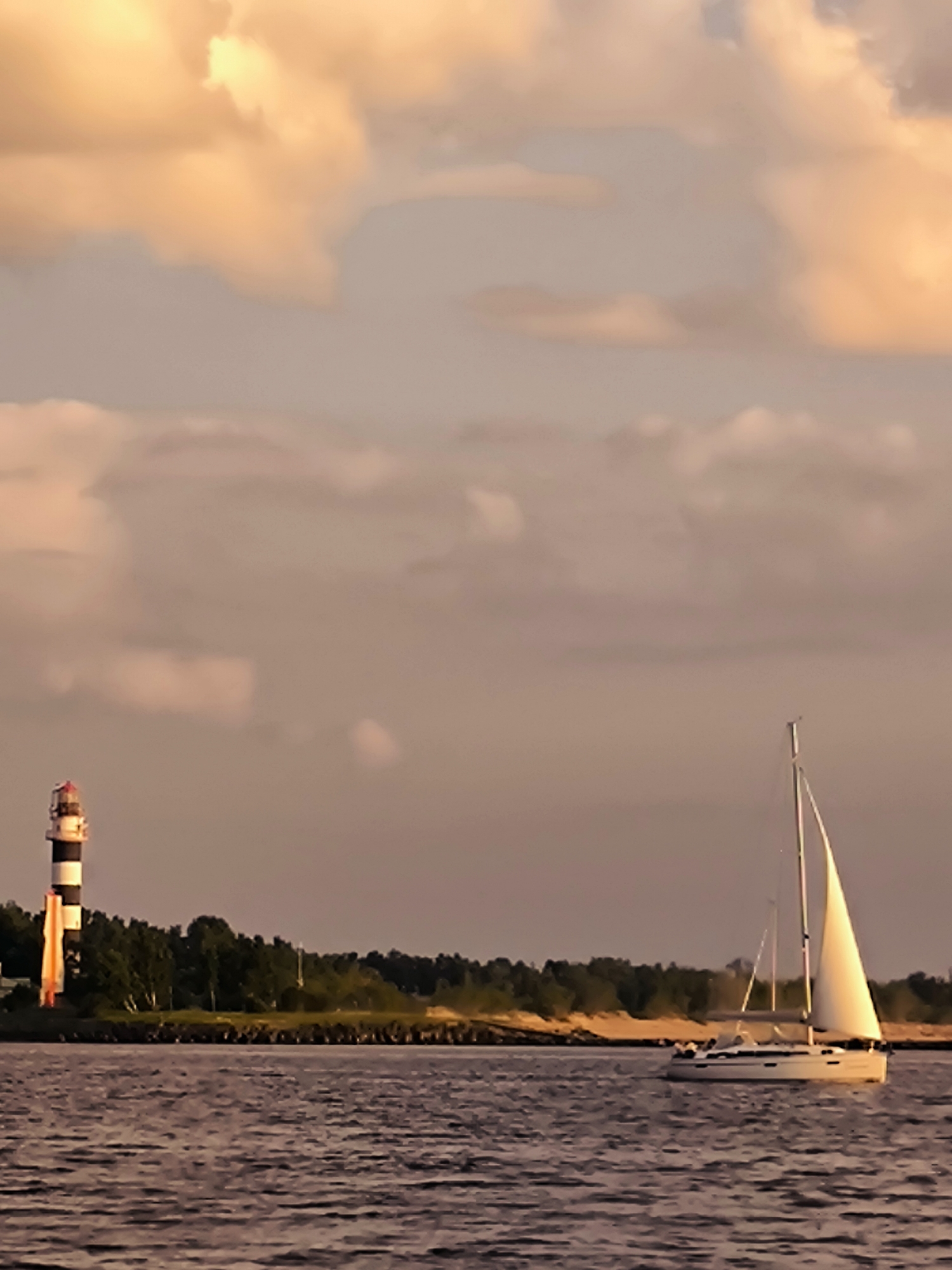 A sailboat with white sails glides across dark, rippling water in front of a tree-lined shore. A striped lighthouse stands on the left side of the image, near the shore. The sky above is filled with soft, orange and pink clouds. The scene appears to be a calm evening or sunset. This image was likely taken in Riga, Latvia, possibly near the Daugava River or the Gulf of Riga.