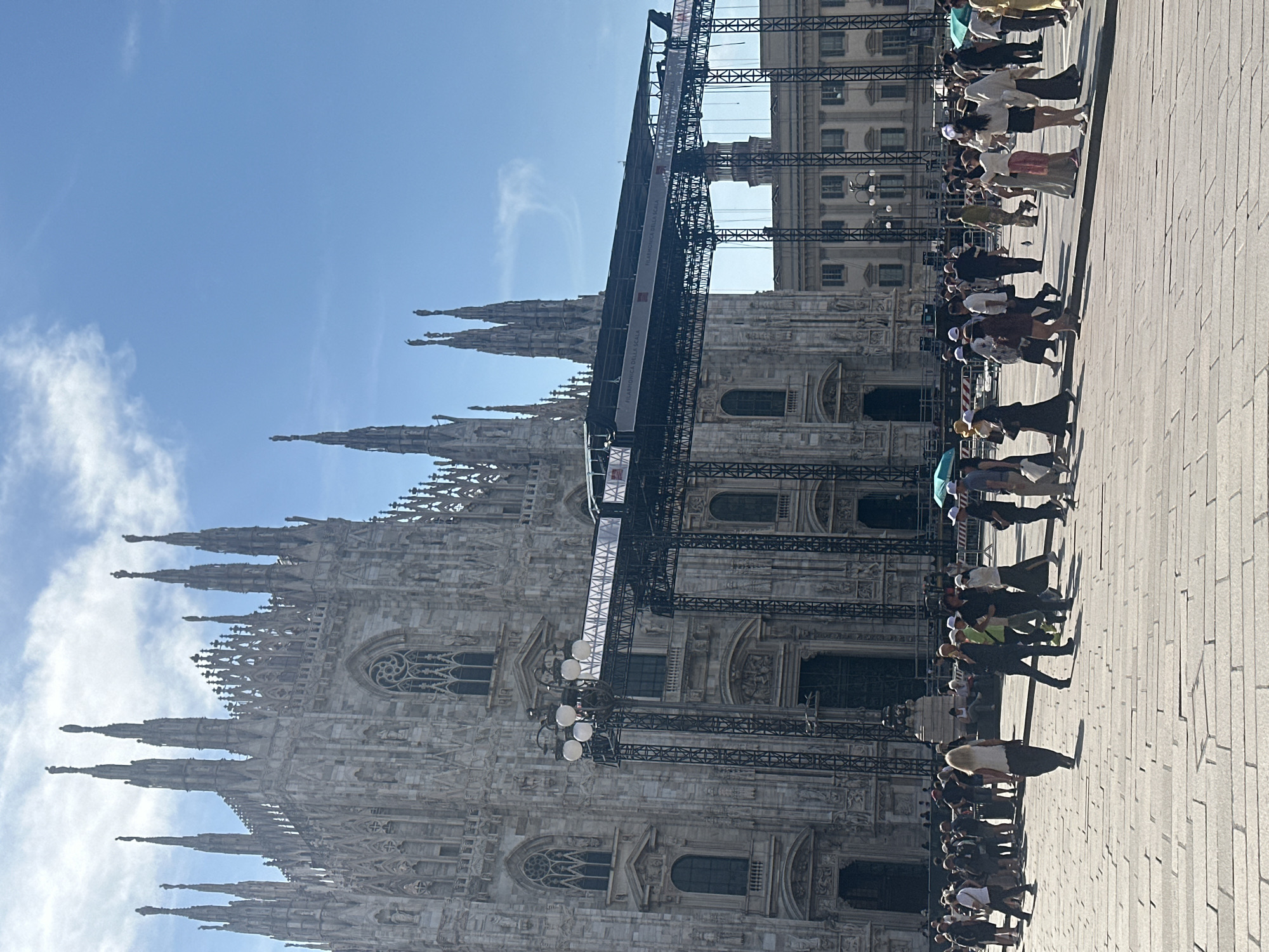 The image shows a view of the Milan Cathedral (Duomo di Milano) in Milan, Italy, with a large temporary stage structure erected in front of its facade. A crowd of people are walking and standing in the piazza in front of the cathedral. The cathedral is made of white marble and features intricate Gothic architecture with spires and statues. The stage is a complex metal framework with a black roof, displaying banners that read "FILARMONICA DELLA SCALA". The sky is clear and blue with some scattered clouds. The ground is paved with large stone tiles. The people in the crowd are dressed in casual summer clothing. Some are looking at the cathedral, while others are engaged in conversation. The overall scene suggests an outdoor event or gathering taking place at a major tourist attraction.