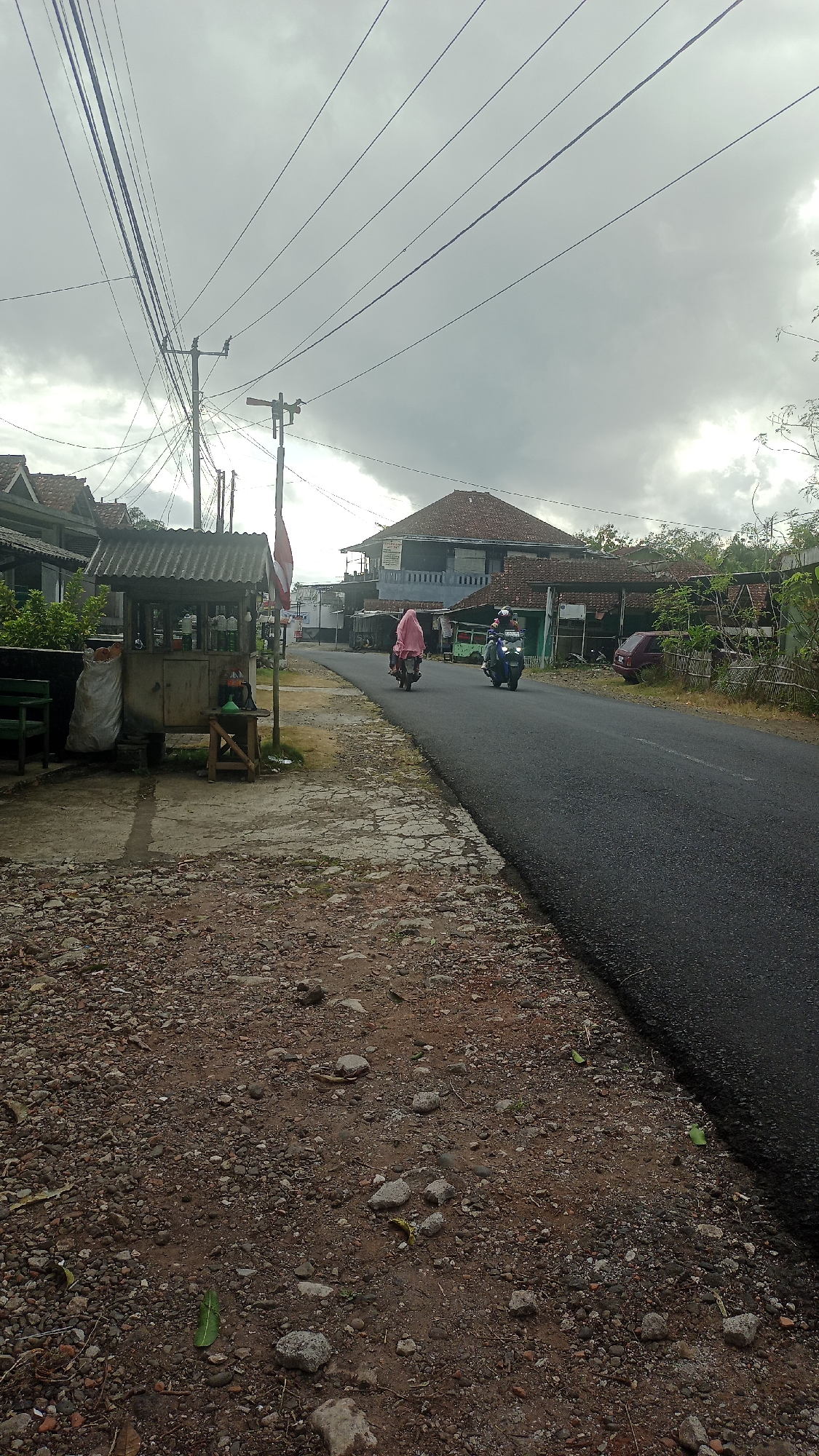 In Karyamandala, Indonesia, a road scene unfolds under a cloudy sky. The foreground is characterized by a rough, unpaved area with scattered gravel and stones. To the left, a small, weathered wooden stall with a corrugated metal roof stands beside the road. Inside the stall, several green bottles, possibly containing beverages or fuel, are visible. A large white sack rests next to the stall, and a green bench is positioned to its left. A wooden stool is placed near the stall.
The road itself is paved and curves slightly to the right. Two individuals are riding motorbikes on the road. The first motorbike, slightly ahead and to the left, carries a person wearing a pink headscarf. The second motorbike, to the right, is ridden by a person wearing a helmet. In the background, houses with traditional tiled roofs line the street, interspersed with trees and vegetation. Utility poles and a complex network of electrical wires crisscross the upper part of the image, contributing to the utilitarian feel of the scene. The overall atmosphere is subdued, likely due to the overcast weather.