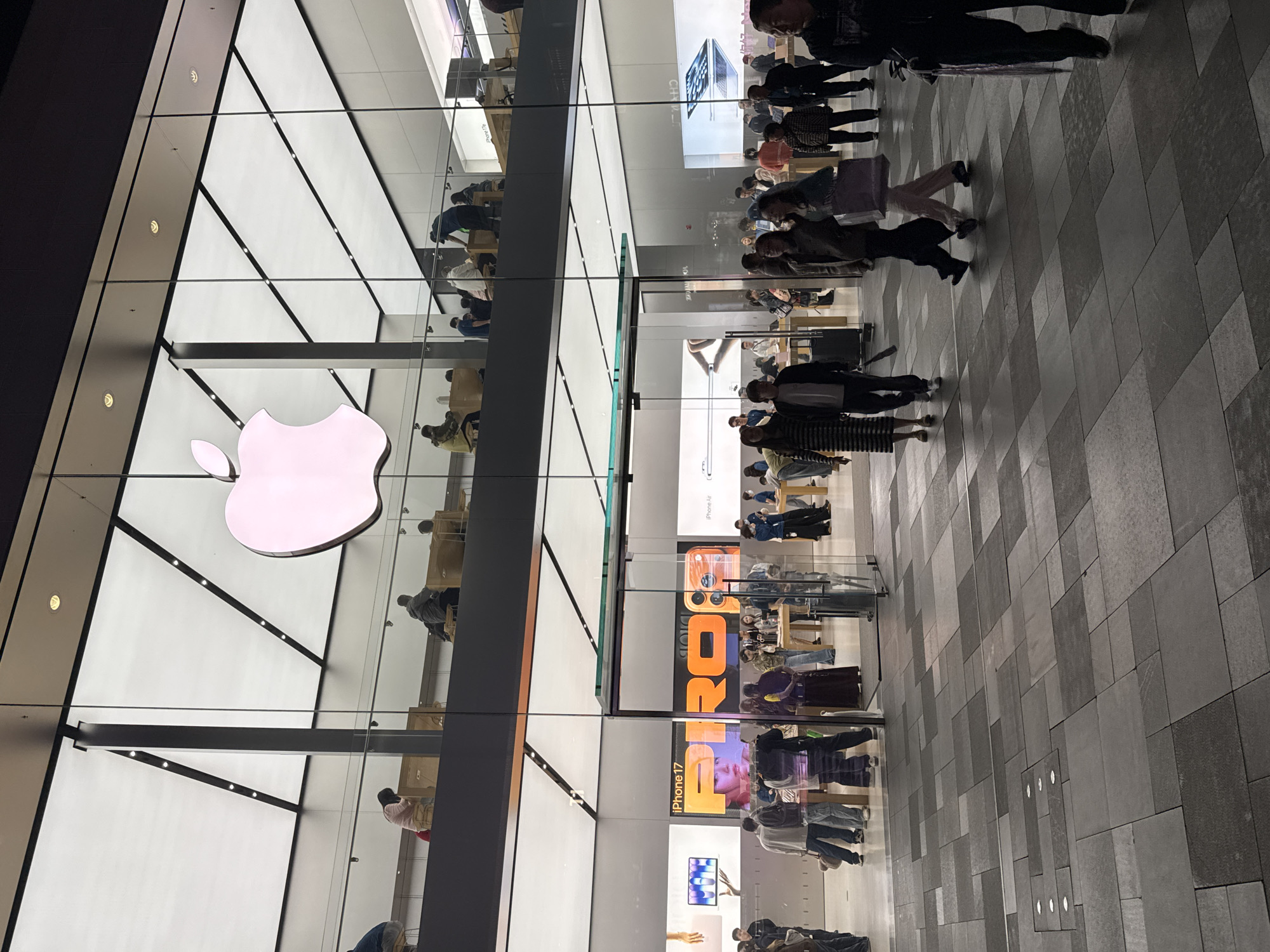 An Apple store is located in the Jinjiang District of China. The store is reflected in the glass facade. Customers are both inside and outside the store. The store is displaying advertisements for the iPhone 17 and the Pro model.