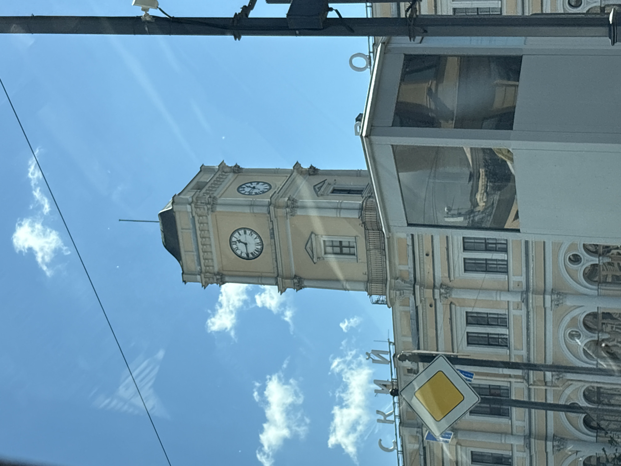 The image shows a bright, sunny day with a clear blue sky and scattered white clouds. In the foreground, a white kiosk with glass windows is visible. To the left of the kiosk, a yellow diamond-shaped traffic sign is mounted on a pole. Behind the kiosk and the sign, a large, pale yellow building with classical architectural features is seen. This building has multiple windows and ornate details. Prominently featured on the building's facade is a tall clock tower with two visible clock faces, each displaying Roman numerals and hands. Above the building's roofline, large white Cyrillic letters spell out "СКИЙ". The overall scene appears to be a street view in Saint Petersburg, Russia, likely depicting a railway station given the architecture and the presence of signage suggesting a transportation hub. There are no people clearly visible in the image.