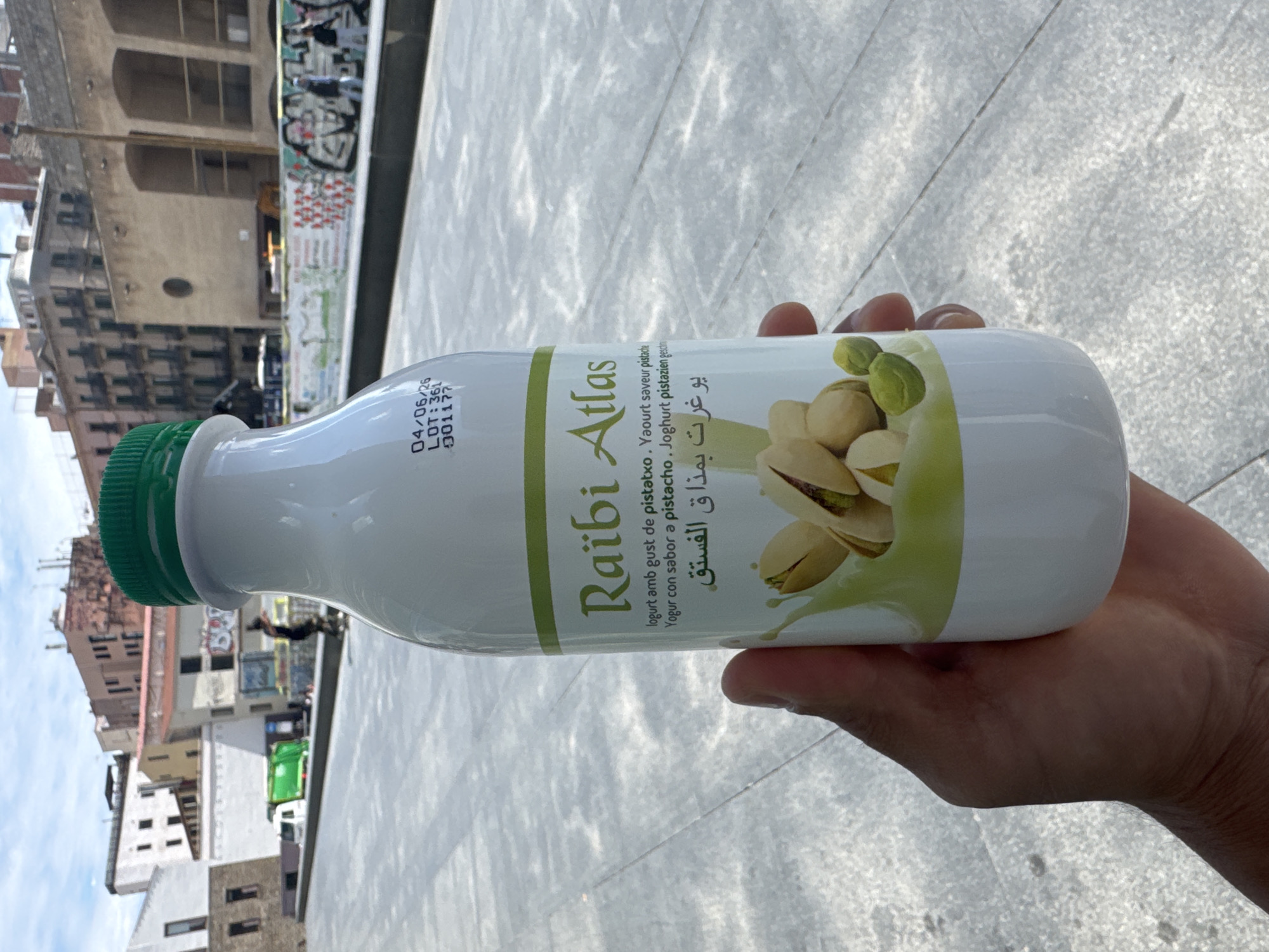 A human hand holds a white bottle of Raïbi Atlas pistachio-flavored yogurt against the backdrop of a Barcelona city scene, Spain. The bottle has a green cap, and the label features pistachios. The yogurt's description is written in multiple languages, including Catalan, French, Spanish, German, and Arabic. The expiration date is printed as "04/06/26" along with the lot number "LOT:361" and the code "001177". In the background are Barcelona city buildings, a street, and a sidewalk.