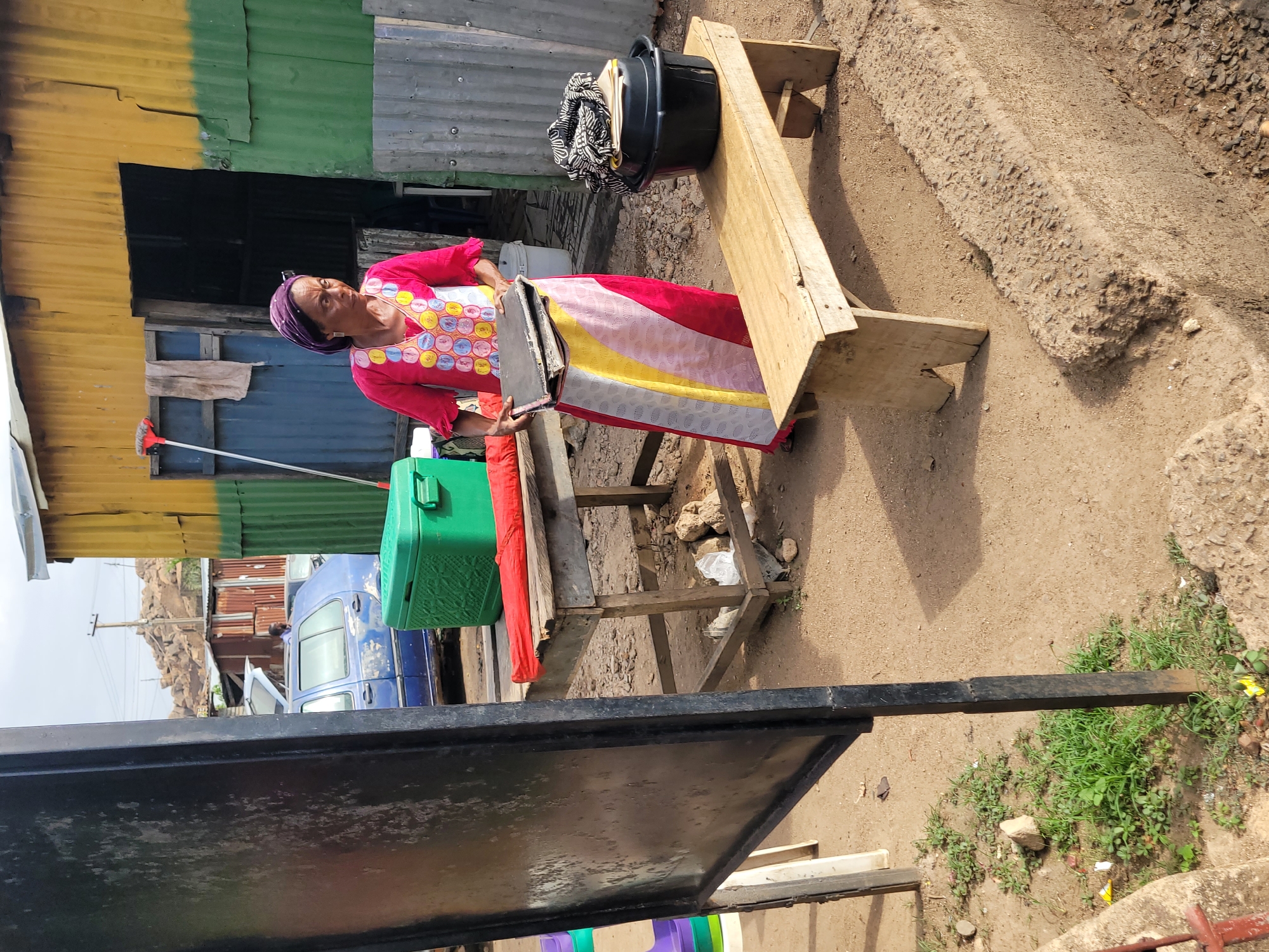 This image captures a vibrant scene in what appears to be a market or residential area in Jos, Nigeria, during daylight hours. The main subject is a woman standing outdoors, dressed in a bright pink long-sleeved top with a colorful, patterned chest area featuring circular designs. She wears a purple headscarf and a long skirt or dress with horizontal stripes of pink, white, yellow, and red. She is holding a flat, dark-colored object, possibly a tray or container, in front of her.
The setting is characterized by informal structures made of corrugated metal sheets, painted in colors like yellow, green, and blue, forming the background. The ground is unpaved dirt, and there's a raised concrete or rocky barrier to the right. To the woman's left, a long-handled broom with red bristles leans against a structure. A bright green cooler or box is visible near her feet. Further back, a blue utility vehicle or truck is parked, and a rocky, arid hill is visible in the distance under a clear sky.
To the right of the woman, there's a makeshift wooden table or stand. On it sits a black plastic tub containing folded fabrics or clothes, possibly for sale. In the foreground, a dark, flat surface, likely part of another stall or barrier, partially obstructs the view. The overall impression is one of daily life in a bustling, informal market or residential zone, with the woman engaged in an everyday activity, possibly trading or organizing goods. No discernible text is visible in the image.