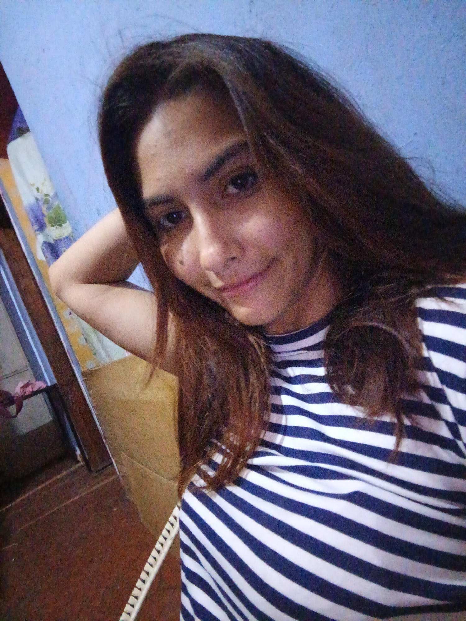 A woman with long brown hair poses for a selfie. She is wearing a striped blue and white shirt. Her left arm is raised and resting behind her head. The background shows a blue wall and a glimpse of furniture, including a wooden frame and a patterned mattress. The flooring appears to be dark. There is no verifiable information to locate the scene in Juan Griego, Venezuela.