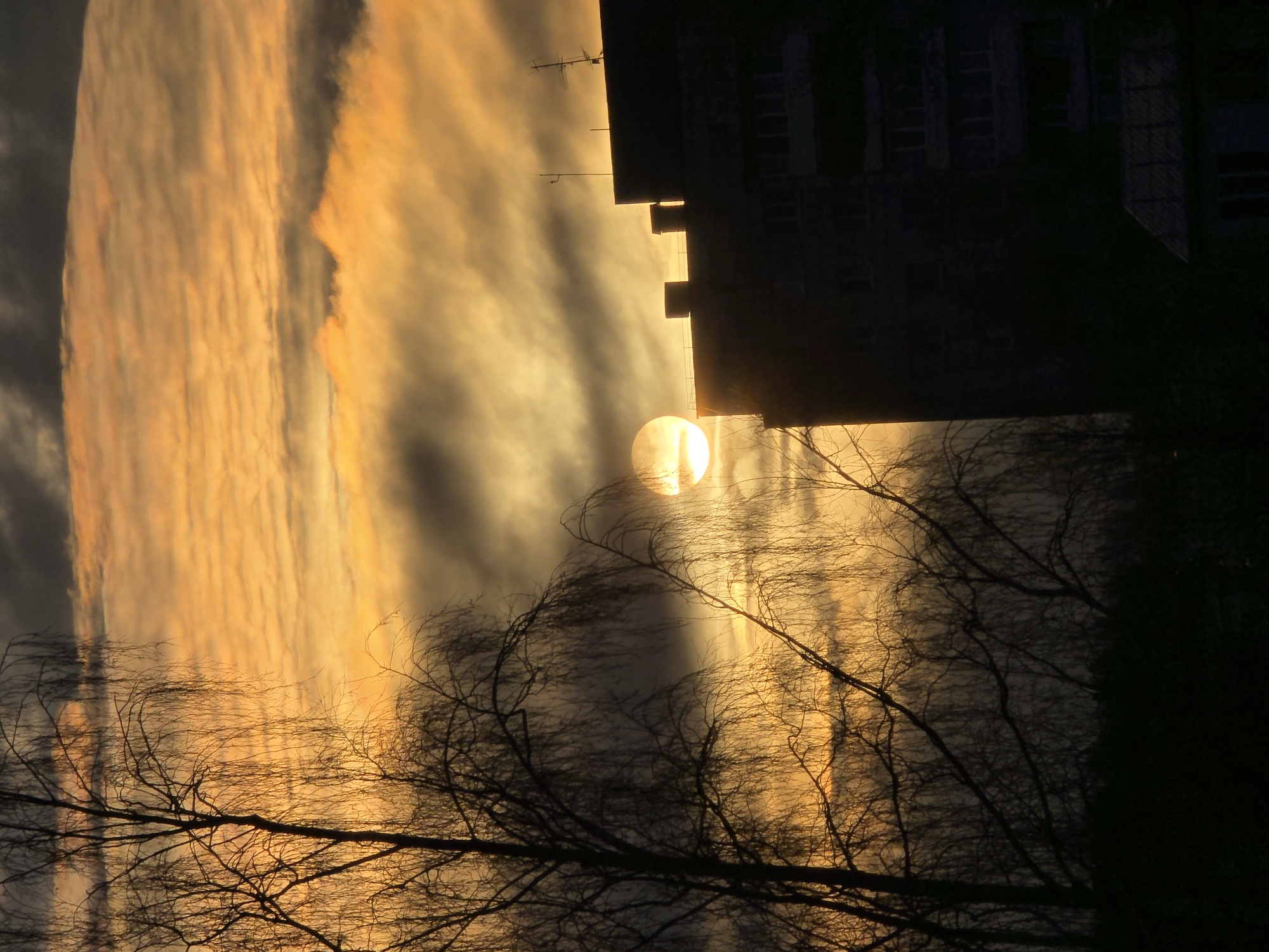 A photograph taken in the city of Unknown, Unknown, captures a low-angle view of the sun partially obscured by a building during what appears to be either sunrise or sunset. The sky displays vertical bands of orange and gray clouds. A dark, silhouetted building stands against the sky, with the sun peeking through a gap in its structure. In the foreground, bare tree branches are silhouetted against the glowing sky.