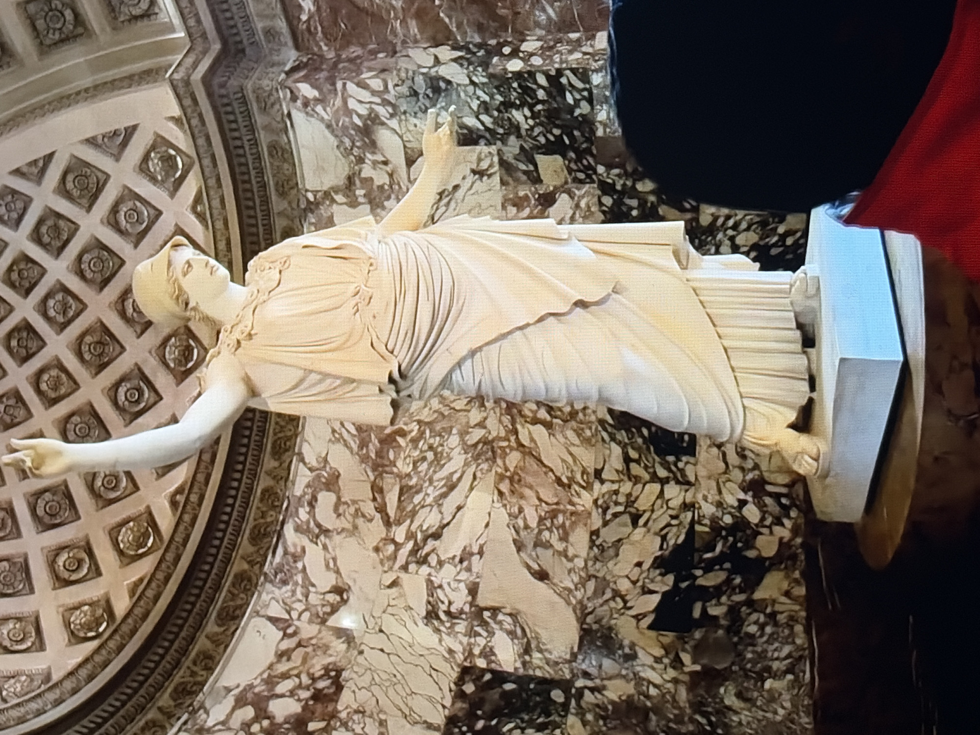 A full shot shows a marble statue of a woman in a classical style, dressed in a draped chiton and a helmet-like headpiece. Her left arm is raised, and her right arm is extended to the side. The statue stands on a light blue rectangular pedestal, which sits on a round wooden base. The background features a marble wall with a pattern of brown and beige veining, and a domed ceiling with ornate, square decorative elements. The bottom right corner of the image shows the back of a person's head, covered in black hair, and a portion of a red garment. The lighting is somewhat dim, suggesting an indoor setting like a museum or gallery.
