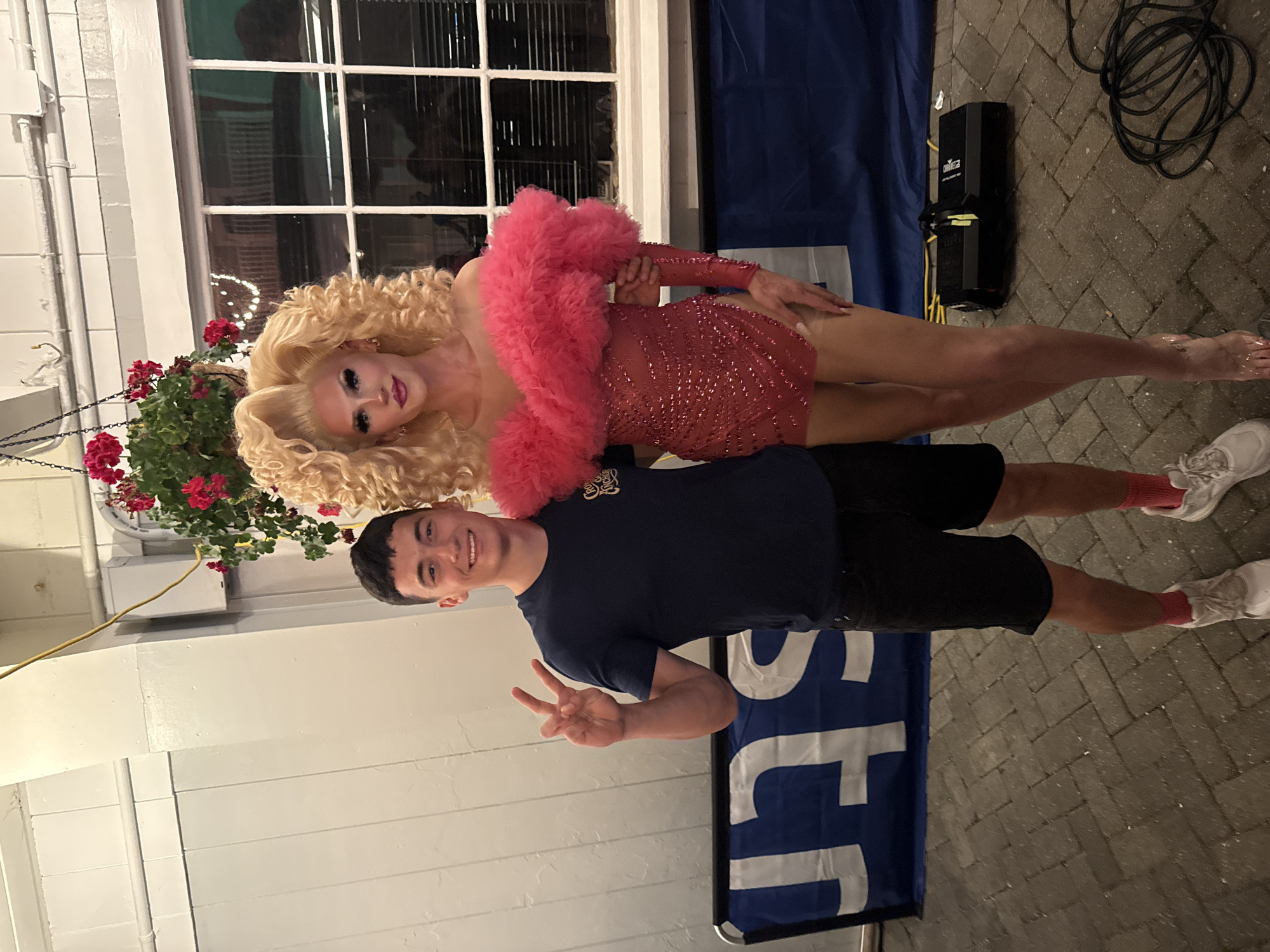 A young man wearing a dark blue t-shirt and black shorts stands to the left of a drag performer in a sparkling red dress with a pink feathery collar and voluminous blonde wig. The man is smiling and holding up two fingers in a peace sign. They are standing on a brick-paved area, likely outdoors. In the background, there is a white wall with hanging plants and a window. A blue banner with white lettering is visible behind them. To the right, a black lighting fixture and coiled cables are on the ground. The scene appears to be in Provincetown, Massachusetts, given the context of drag performers and the likely presence of a venue associated with such entertainment.