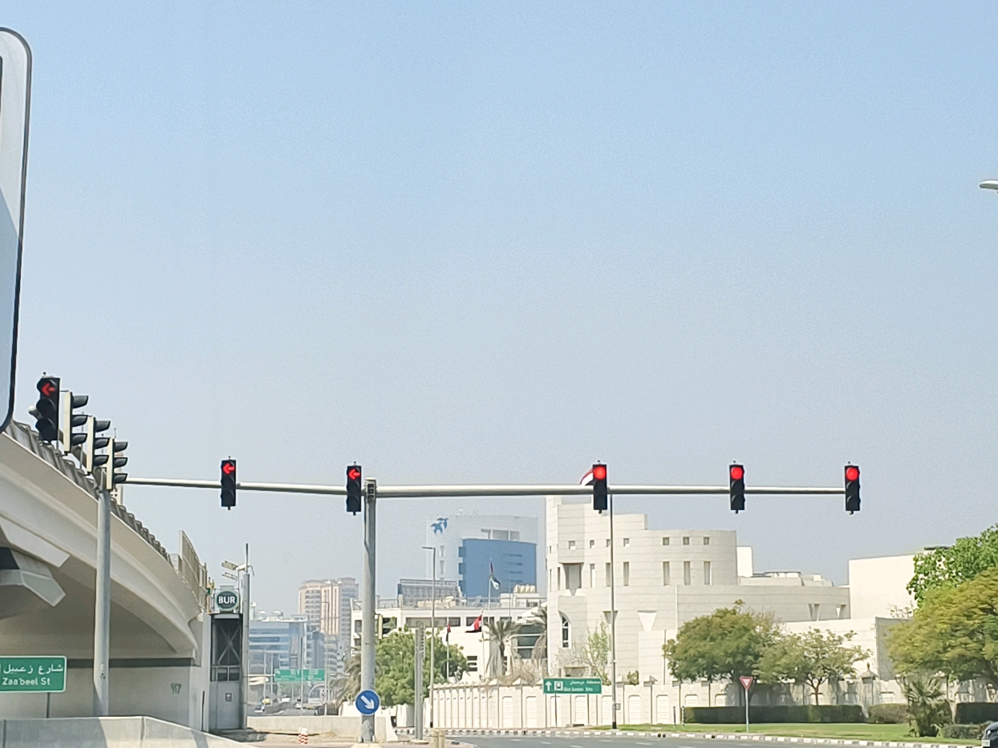 This image captures a street scene in Dubai, United Arab Emirates, likely during daylight hours due to the clear sky and bright lighting. The primary focus is on a series of traffic lights, all displaying red signals, indicating a stop for vehicles. The traffic lights are mounted on horizontal poles spanning across the road.
In the foreground, on the left, a portion of an elevated highway or bridge structure is visible, painted in white. Adjacent to this, a green street sign reads "Zaa'beel St" in English and Arabic script, identifying the location. A smaller sign with the letters "BUR" is also present.
The background reveals modern city architecture, characterized by multi-story buildings with clean lines and light-colored facades. Some buildings have a more institutional or governmental appearance, with flags visible on some structures. Trees and landscaping are interspersed between the buildings, suggesting a well-maintained urban environment. The sky is a pale blue, with no visible clouds, indicating fair weather conditions. There are no people or vehicles clearly discernible in the immediate scene. The overall impression is of a busy intersection in a contemporary urban setting within Dubai.