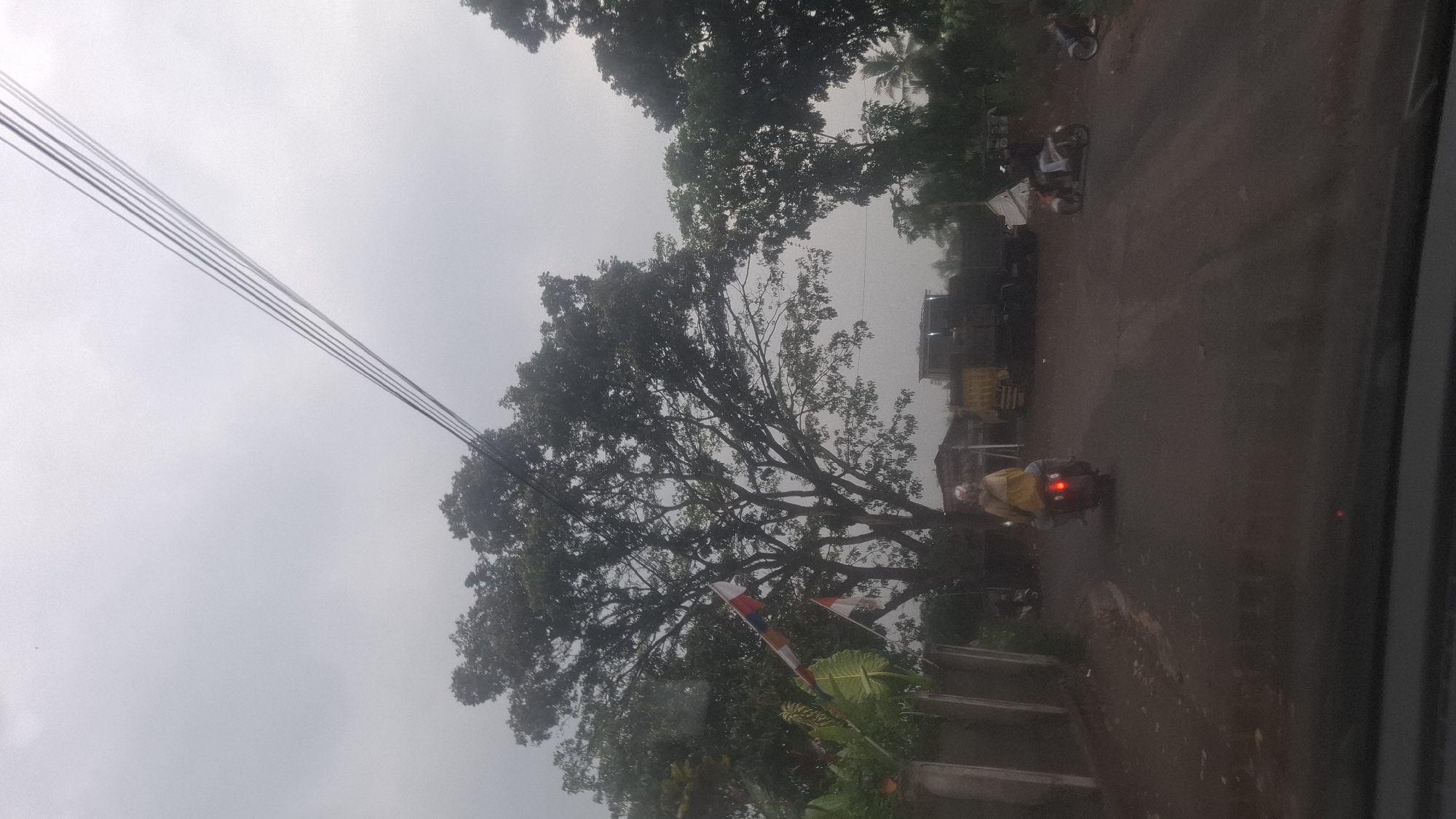 In Cibinong Hilir, Indonesia, on an overcast day, a rural dirt road stretches into the distance. A person wearing a yellow jacket rides a motorcycle away from the camera, their brake light illuminated. To the left, a tall tree with lush green leaves dominates the frame, adorned with two Indonesian flags. Further down the road, several trucks are parked, one with a yellow cargo bed. Another motorcycle is visible on the right side of the road, parked. The scene is framed by electrical wires spanning the sky and a concrete wall along the left edge of the road.