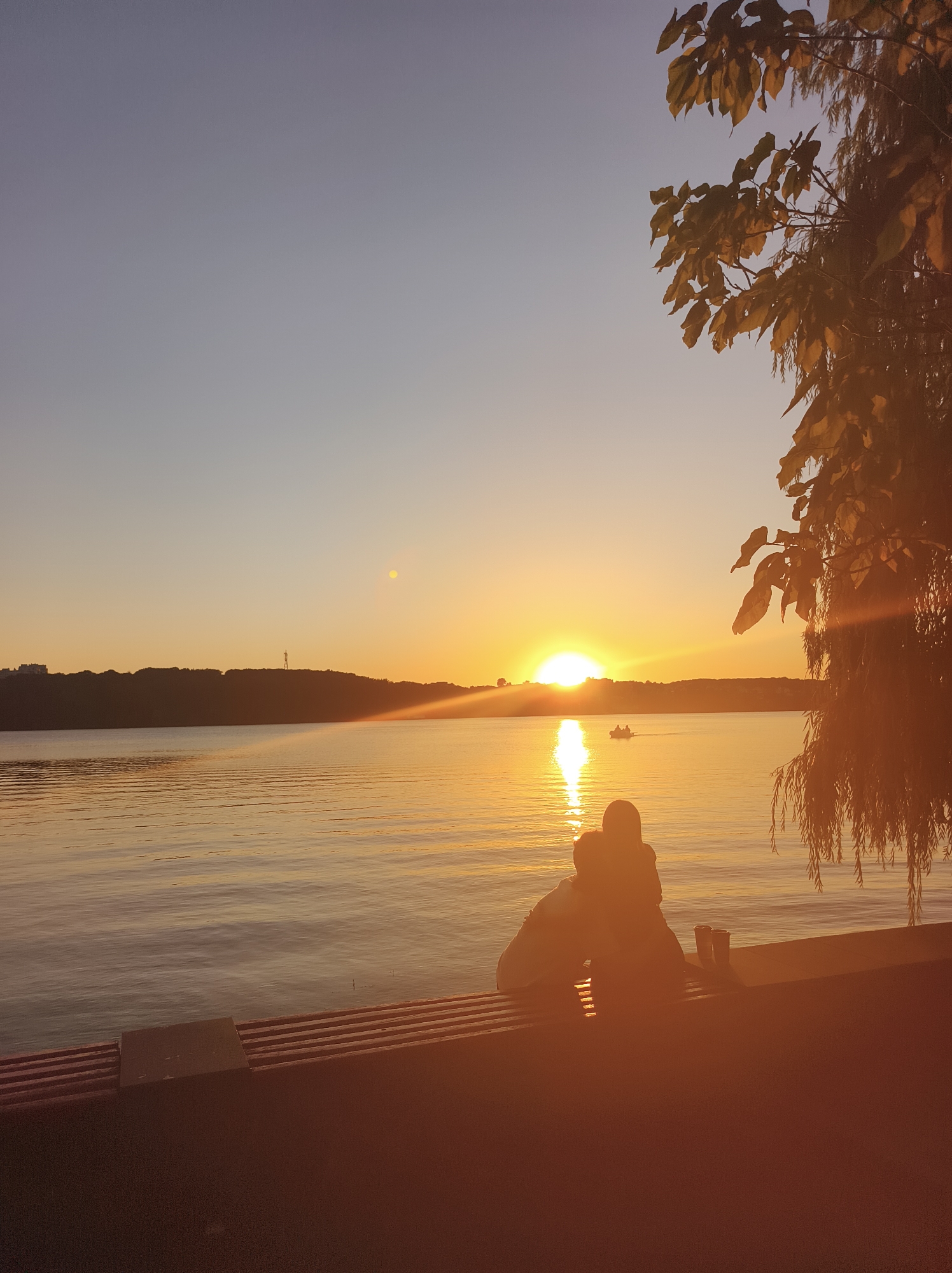 The image depicts a scenic sunset over the Ternopil lake in Ternopil, Ukraine. In the foreground, two individuals are seated on a bench, silhouetted against the warm glow of the setting sun. They are facing the lake, appearing to be in an embrace or close proximity. To the right of the couple, a willow tree with drooping branches partially obscures the view, its leaves catching the golden light. The lake stretches out before them, its surface rippling gently and reflecting the vibrant colors of the sunset. In the distance, a tree-lined shore forms a dark silhouette against the bright horizon. A small boat with two people is visible on the water, adding a sense of scale and activity to the tranquil scene. The sky transitions from a soft blue at the top to a warm orange and yellow hue near the horizon, with the sun positioned low and casting a brilliant reflection on the water. The overall atmosphere is one of peacefulness and natural beauty.