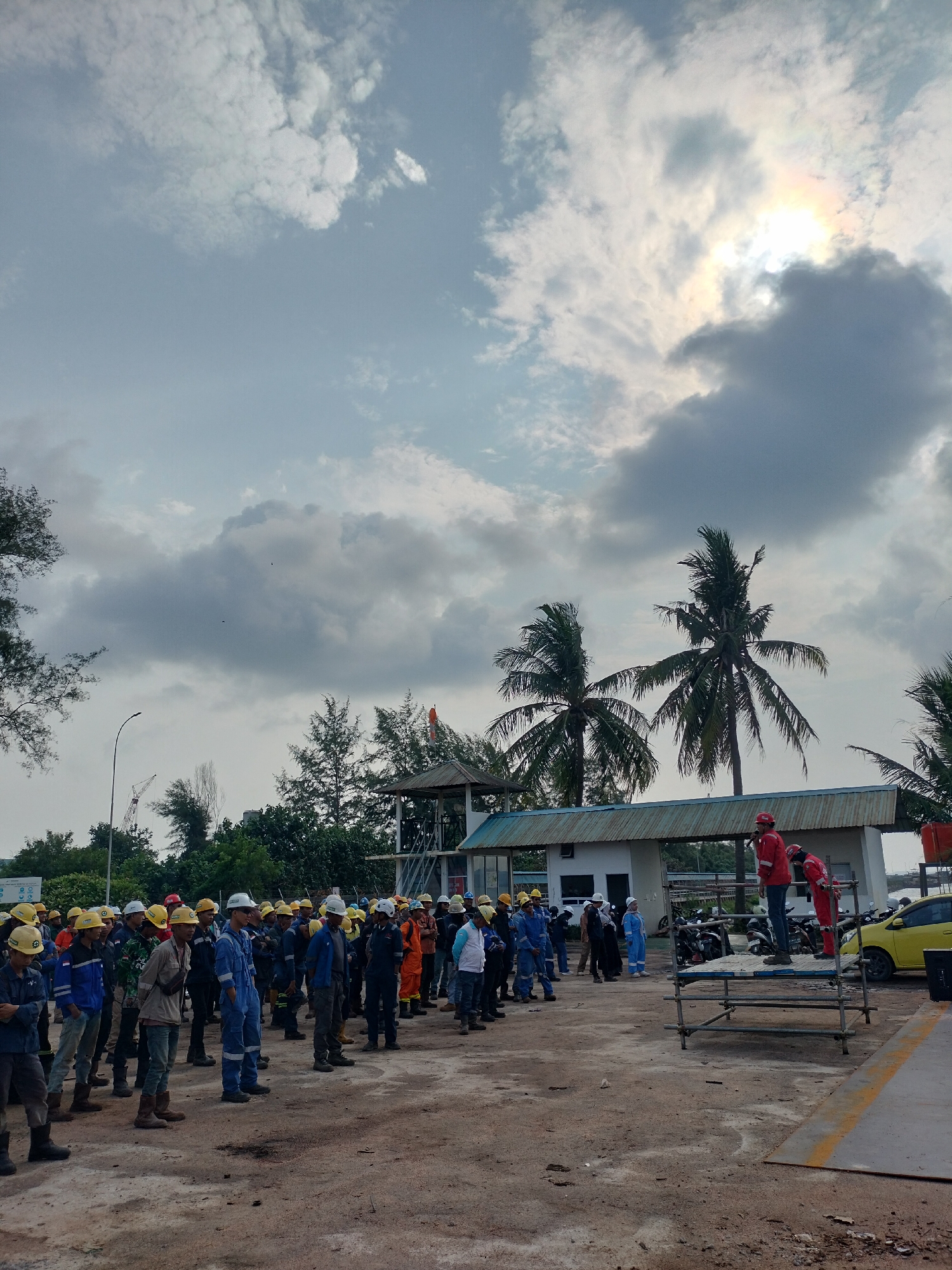 The image depicts a group of workers, predominantly male, gathered outdoors in what appears to be an industrial or construction site. Many are wearing hard hats and work uniforms, suggesting a safety briefing or pre-work assembly. The environment features a dirt ground, a low-rise building with a corrugated metal roof, palm trees, and a cloudy sky with patches of sunlight breaking through, creating a hazy atmospheric effect. In the background, a yellow car is visible, along with several motorcycles parked near the building. The overall scene suggests an active work environment in a tropical or subtropical region, potentially within the city of Batam, Indonesia, given the presence of palm trees and the general architectural style of the buildings and infrastructure. There are two individuals in red safety vests standing on a temporary scaffolding structure, possibly addressing the assembled group. The lighting indicates it is daytime, likely late afternoon due to the low angle of the sun and the diffused light.