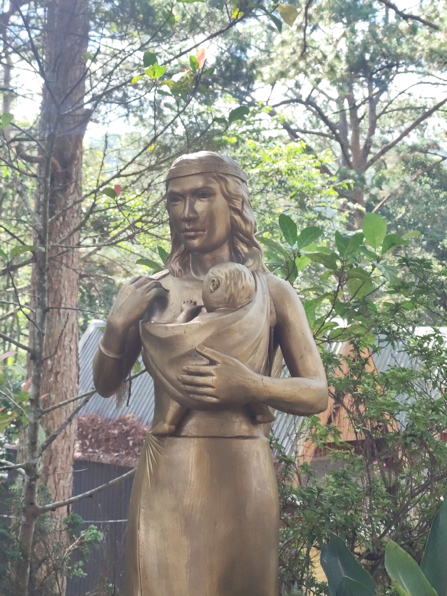 A bronze statue of a woman carrying a baby is positioned outdoors in front of a backdrop of trees and foliage. The woman is wearing a headpiece and her hair is long and wavy. She is holding the baby wrapped in a cloth against her chest. The statue is golden in color. The background consists of various trees with sunlight filtering through the branches, as well as some green leafy plants. In the distance, a building with a corrugated metal roof is visible. This statue is located at the Da Lat Flower Park in Da Lat, Vietnam.
