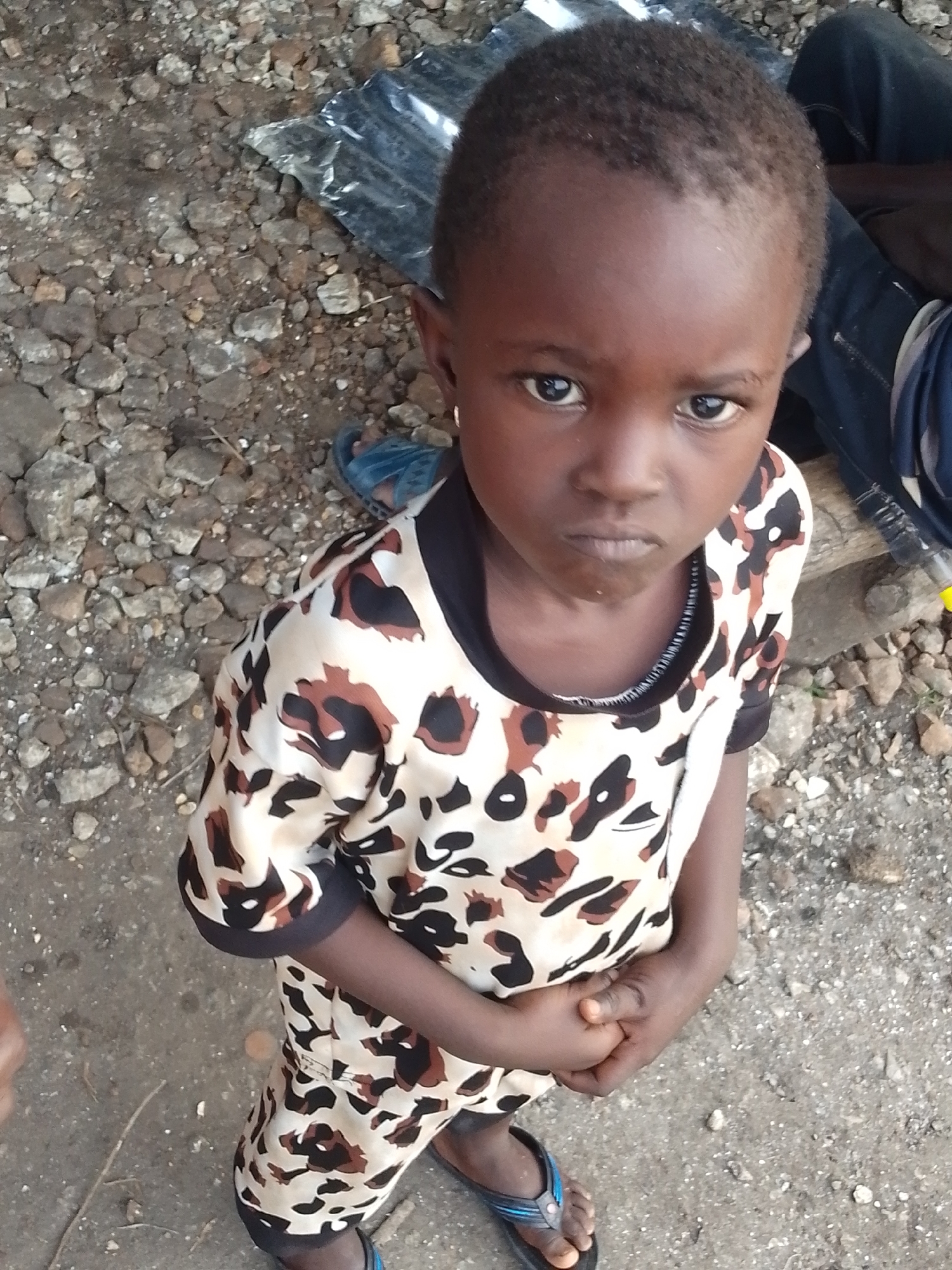 A young Black child, likely a girl based on facial features, stands in a seated or squatting position outdoors. The child has dark skin and short, dark hair. She is wearing a short-sleeved top with a leopard print pattern in black, brown, and beige. The child's hands are clasped together in front of her. She is wearing dark flip-flops on her feet. The ground beneath her appears to be dirt and gravel, with some scattered rocks and debris. In the background, to the right, a person's legs are visible, wearing dark blue jeans. There is also a piece of dark plastic sheeting and a wooden plank or log in the background. The scene is consistent with an outdoor environment in a rural or informal settlement, such as what might be found in Otulu, Nigeria.
