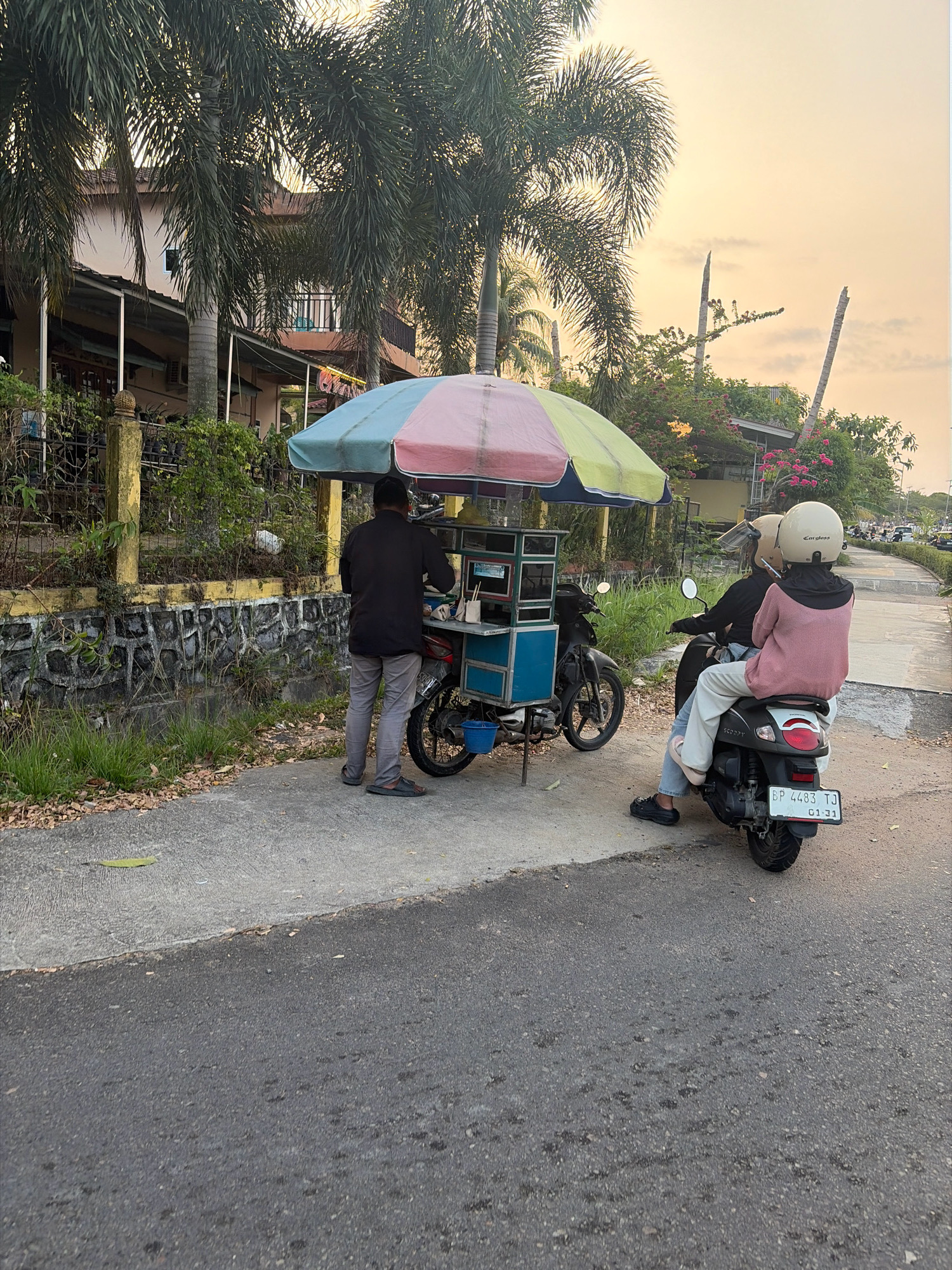 In Tanjung Pinang, Indonesia, at approximately 6:00 PM on a cloudy day, a street vendor operates a food cart under a colorful umbrella. The cart is attached to a motorcycle. A man, dressed in a dark long-sleeved shirt and grey pants, stands behind the cart, appearing to be serving customers. Two individuals, one driving and one a passenger, are seated on another motorcycle parked near the food cart, facing away from the camera. The passenger wears a pink long-sleeved shirt and white pants, while the driver is in a dark shirt. Both wear helmets. The scene is located on the side of a paved road with a sidewalk. Palm trees and vegetation are visible behind a stone wall bordering a property. The sky is overcast with a warm, golden hue, suggesting sunset. The license plate on the motorcycle with two people reads "BP 4483 TJ 01-31".