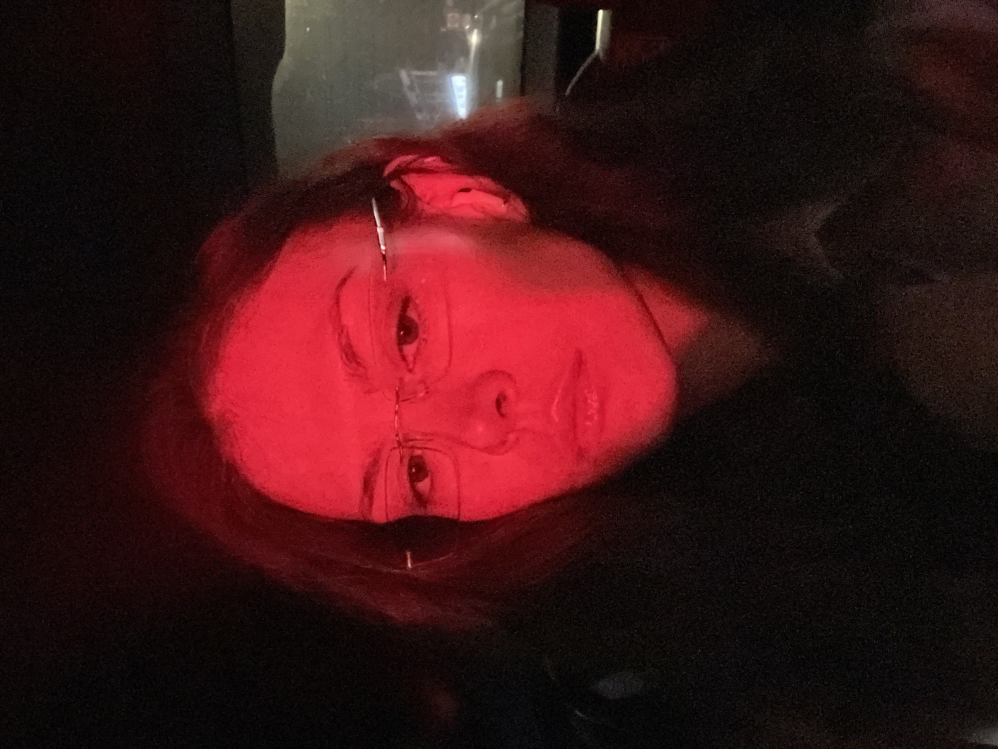 A close-up portrait features an individual's face, strongly illuminated by a vibrant red light. The person's head is tilted due to the image being rotated clockwise. They wear glasses, visible across the bridge of their nose and around their eyes, with the frames reflecting some of the intense red light. Their eyes are open and directed towards the viewer, with a neutral to slightly thoughtful expression. Dark hair frames the face, blending into the surrounding darkness. The red light casts dramatic shadows and highlights, emphasizing facial contours and creating a striking, monochromatic effect on the skin.
The background is predominantly dark, almost black, with a grainy texture suggesting low-light conditions. In the upper portion of the original image and some crops, faint, blurry, and out-of-focus lights are discernible. These distant light sources suggest an urban environment at night, possibly viewed through a window, or ambient lighting within a dimly lit interior. There is no visible text. The overall atmosphere is moody and intense, characteristic of night photography in a setting like London, United Kingdom.