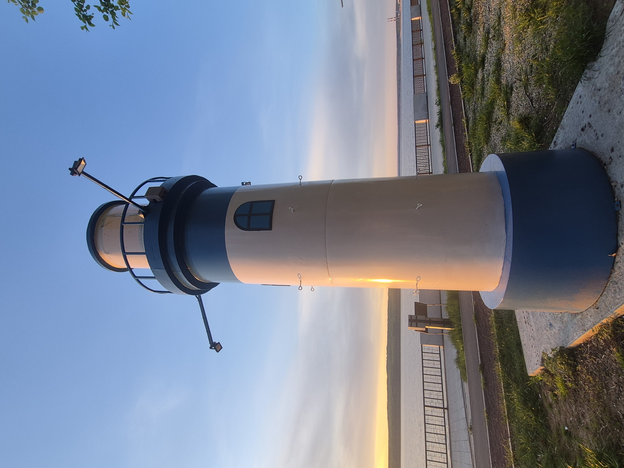 A vertical shot shows a lighthouse at sunset on the embankment in the city of Chaikovsky, Russia. The lighthouse has a blue base, a beige body, and a blue top. The sky is blue, but near the horizon, it is yellow, representing the sunset. In the background is the embankment, a body of water, and a strip of land.