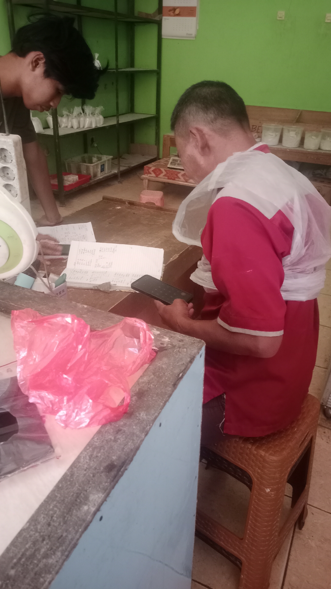 In a room with green walls and shelves, two men are present. The man on the left, wearing a dark shirt, is leaning over a counter with a white object that appears to be an electrical socket and a scale. In front of him are papers and a red plastic bag. The man on the right, wearing a red shirt and a white plastic cover over his shoulders, is seated on a brown plastic stool. He is holding a black smartphone in his right hand and looking down at it. Behind the man on the right, a wooden bench holds several small white containers. The scene suggests a workspace or storage area, possibly in Karyamandala, Indonesia, given the presence of Indonesian currency visible on the papers in front of the man on the left.