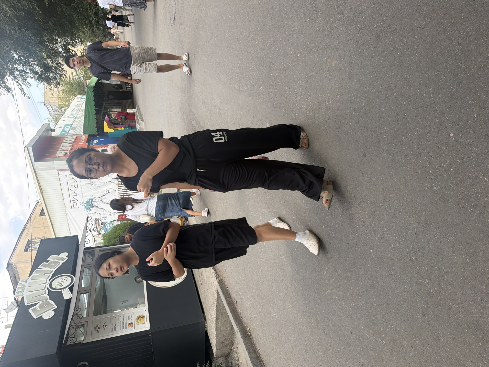 In this full-body shot taken outdoors, two young Asian women are walking on a paved city street. The woman in the foreground on the right is wearing black, wide-leg pants with "04" and some text in white on the left leg, and a black t-shirt. She has dark hair and is wearing glasses and sandals. The woman on the left is wearing black shorts and a black t-shirt, with white socks and white slip-on shoes. She has dark hair and appears to be looking to her left.
In the background, a young Asian man in a dark t-shirt and light shorts with white slides is walking away from the camera. Other pedestrians, buildings, trees, and an outdoor food stall are visible in the background, suggesting a commercial or pedestrian area within a city. The stall has signage in Cyrillic script and an image of pancakes. The sky is overcast.