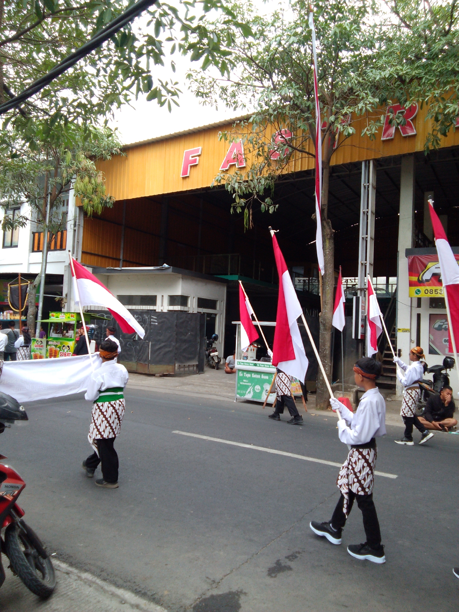 In the city of Trenggalek, Indonesia, on a street, several young individuals are participating in what appears to be a parade or cultural event, walking on the asphalt road. They are dressed in traditional Indonesian attire, featuring white shirts, batik-patterned sarongs or wraps around their waists, and headbands. Many of them are carrying Indonesian flags, which are red and white, along with a long white banner that stretches across the foreground. In the background, there is a building with a yellow facade and large red letters that spell out "FA." Trees with green leaves partially obscure the building. There are also street vendors or stalls visible, one with a green and white sign. A red motorcycle is parked on the left side of the road. Another person is sitting on the side of the road, and a few other individuals are standing around. The scene suggests a public gathering or celebration.