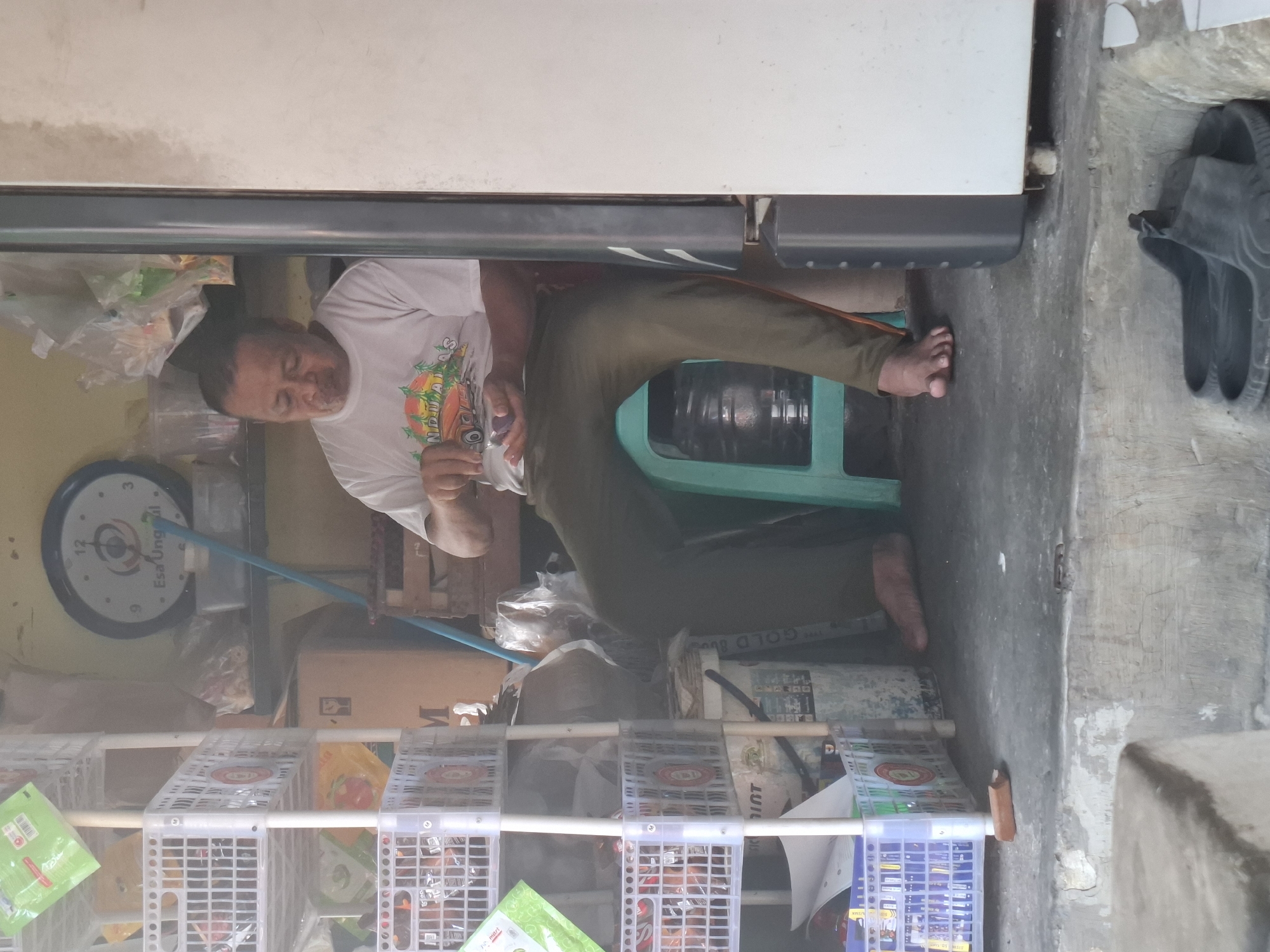 A middle-aged Indonesian man sits on a teal plastic stool in a cluttered, dimly lit shop or storage area, possibly in West Jakarta. He wears a white t-shirt with a graphic print and olive green pants. His gaze is directed downwards as he holds a small, dark object, perhaps candy or a pill, in his right hand. His left hand is also near his mouth, and his facial expression suggests concentration or mild contemplation. The environment is filled with stacked white plastic crates containing various packaged goods and bags. In the background, a large circular clock with a blue frame is mounted on the wall. A blue pole leans against the wall near the clock. The floor is concrete, and a large, light-colored appliance, possibly a refrigerator, is partially visible to the right. A pair of black flip-flops rests on a step in the foreground. The scene conveys a sense of everyday life within a commercial or storage space.