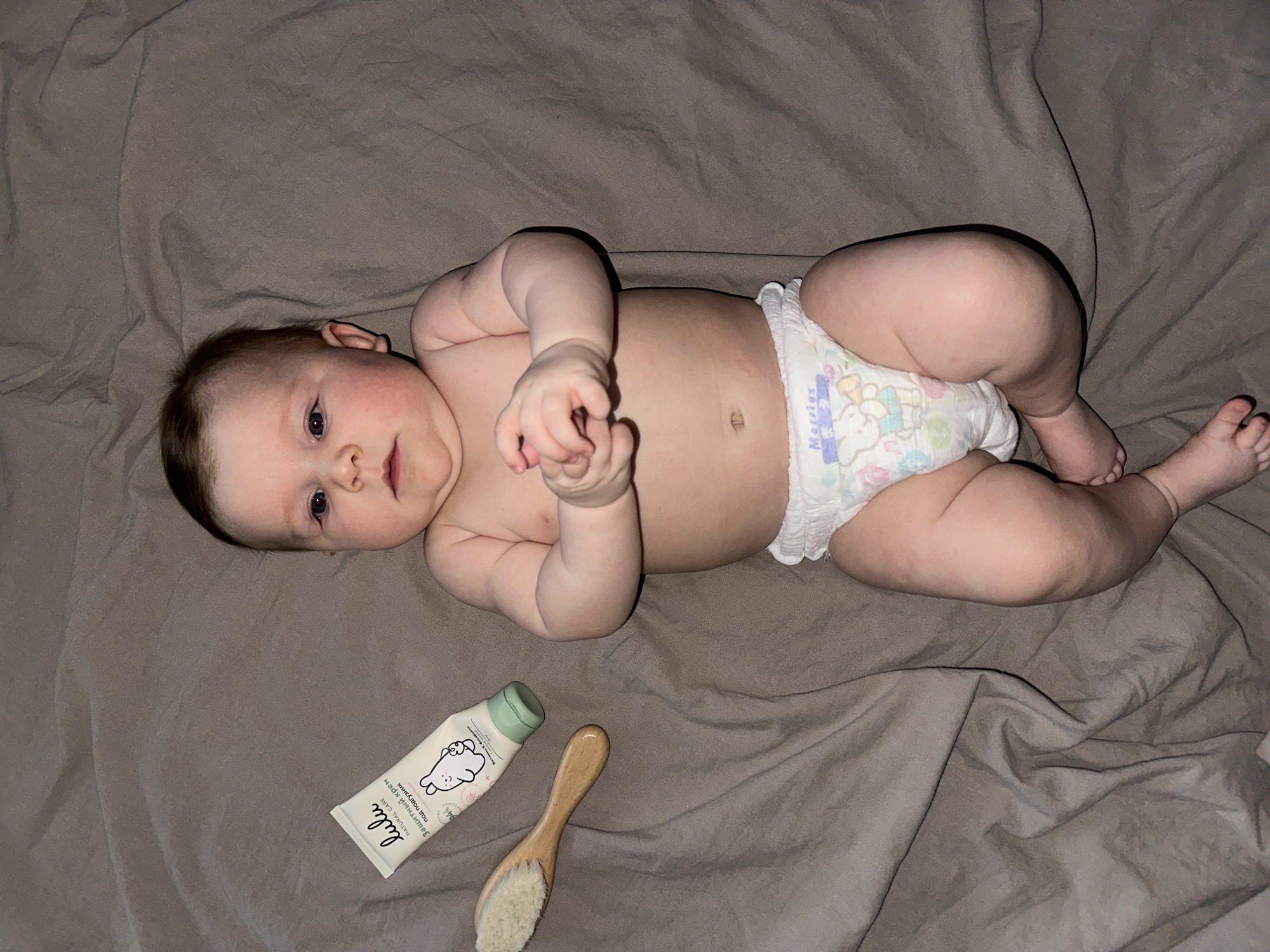 A baby, wearing a diaper, lies on a gray blanket. Beside the baby, a tube of Lulu Natural Care cream and a wooden brush are visible. The location is Chaikovsky, Russia.