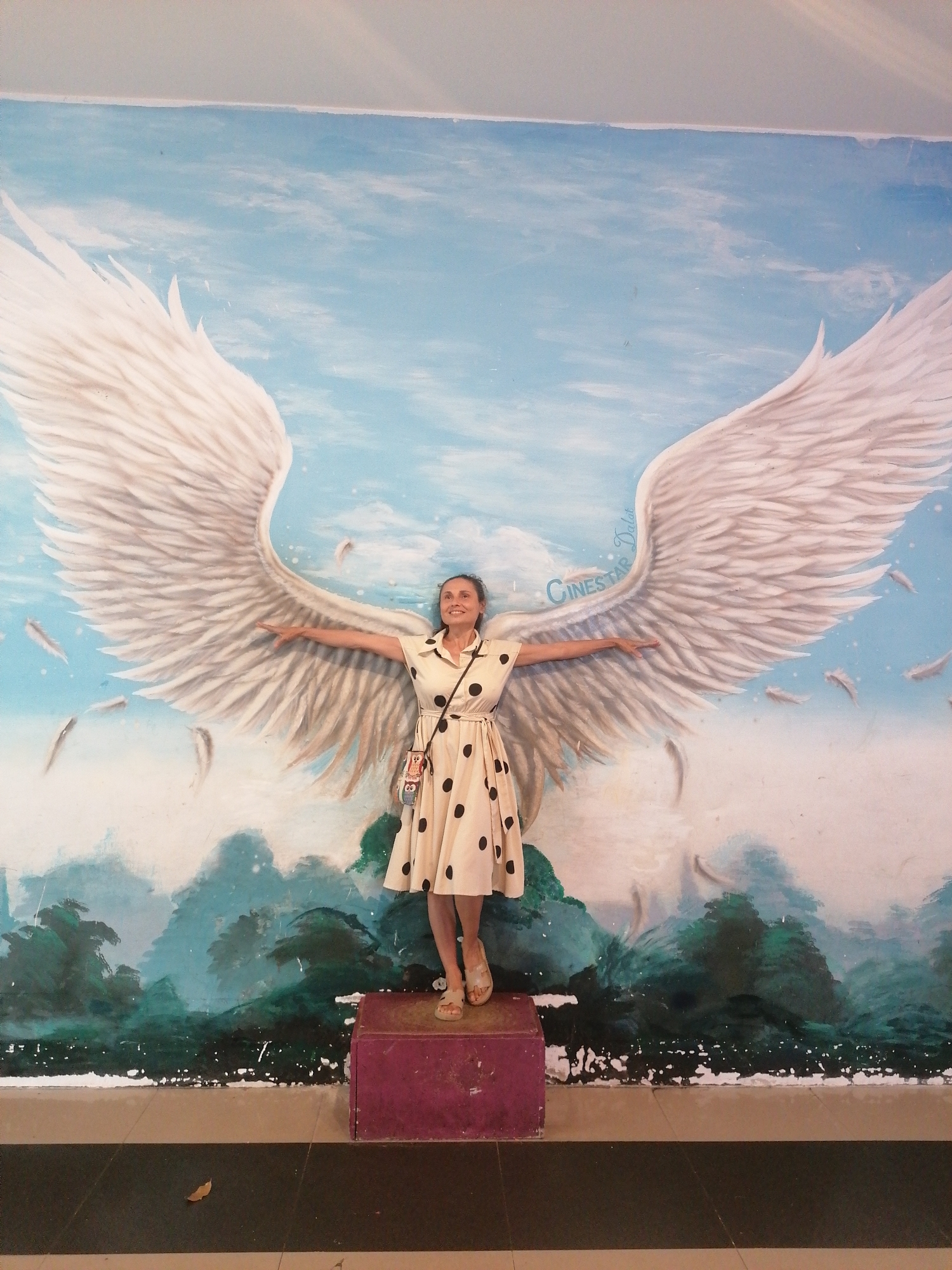 The image captures a woman posing in front of a painted mural, identified by text as "CINESTAR Dalat," indicating a location in Da Lat, Vietnam. The woman stands centrally, smiling with her head slightly turned to her right and upwards. She wears a cream or beige short-sleeved dress with black polka dots and a collar, cinched at the waist. A small, multicolored owl-themed crossbody bag hangs from her left shoulder. She stands on a low, rectangular, dark pink platform. Her arms are extended horizontally to her sides, palms open, aligning with large white feathered wings painted on the mural behind her. The mural depicts a light blue sky with white clouds in the upper half and stylized green and teal mountains or foliage in the lower half. Several individual white feathers are painted floating in the sky around the wings. The floor in the foreground consists of large square tiles in alternating black and light brown stripes, with a single brown leaf resting on a black tile in the lower-left corner. A narrow strip of a light grey ceiling or upper wall is visible at the top of the frame.
