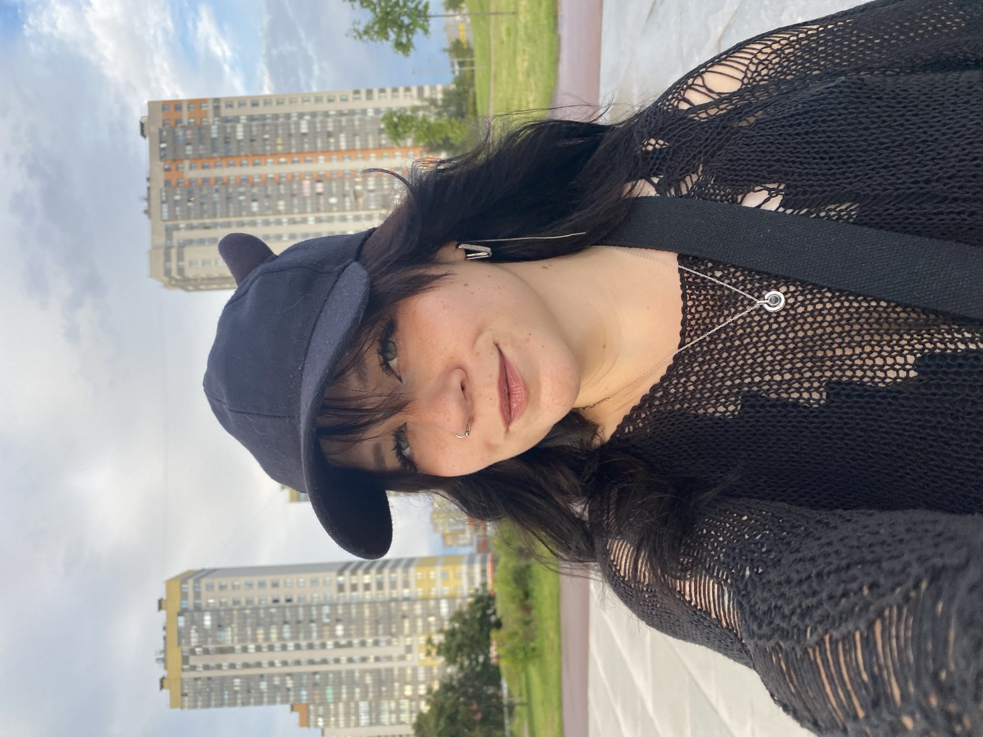 A young person with dark hair, wearing a black baseball cap with ear details, a ripped black knit sweater, and a silver necklace with a circular pendant, takes a selfie outdoors. They are wearing a nose ring and large hoop earrings. In the background, two tall, modern apartment buildings are visible, along with a grassy park area and a cloudy sky. This scene is located in the Moskovsky District of Saint Petersburg, Russia, likely near one of the large residential complexes.