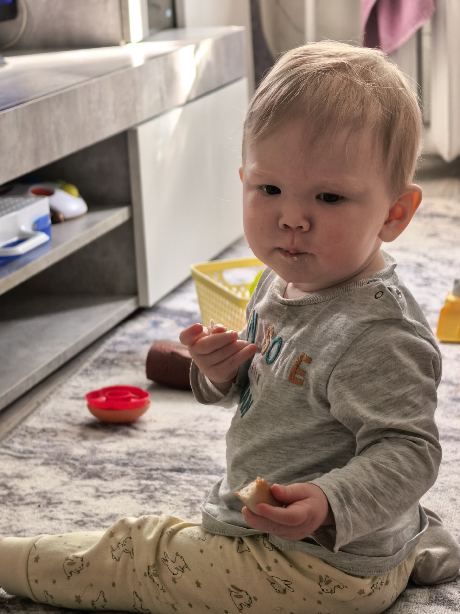 An infant, approximately 1-2 years old, with light blonde hair, sits on a light-colored patterned rug in an indoor domestic setting. The child is dressed in a grey long-sleeve top embroidered with partial letters 'AWESO M E' and yellow pants patterned with dark animals and stars. They are focused, holding a small piece of light-colored food, possibly bread or a biscuit, in their right hand, with a few crumbs visible around their mouth. The background on the left features a low, grey, concrete-effect cabinet with open shelves, displaying a white and blue box and a white and yellow toy car. Strong natural light creates a diagonal highlight on the cabinet's top surface. On the right, a yellow plastic basket, a red and orange toy bowl, and a partially visible yellow bottle are on the rug. A folded pink towel hangs in the upper right background.