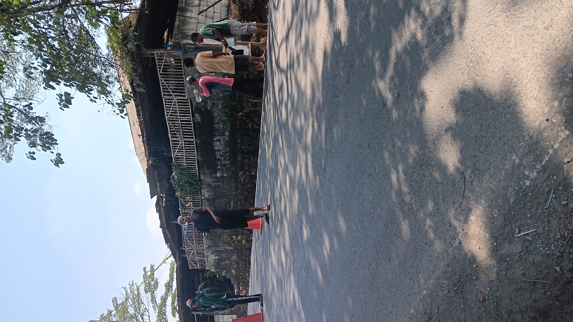 In Jonggol, Indonesia, on a sunny day, several men are seen standing on the side of a road. The road is paved with asphalt and has scattered debris. In the background, a row of buildings with corrugated metal roofs is visible, separated from the road by a stone wall and a wooden fence. Trees with green foliage provide shade on the road, casting dappled shadows.
One man, wearing a dark t-shirt and shorts, stands near the center of the frame, facing forward and appearing to be smoking. To his left, another man in a dark green jacket and cap stands near an orange traffic cone. Further to the right, a man in a pink and grey shirt is drinking from a clear plastic bottle. Next to him, a man in a tan shirt and shorts is standing near a white container on a stand. To the far right, a man in a green shirt and light-colored pants is also present, with his hands near the white container. The overall scene suggests a casual outdoor gathering or a brief pause during work.