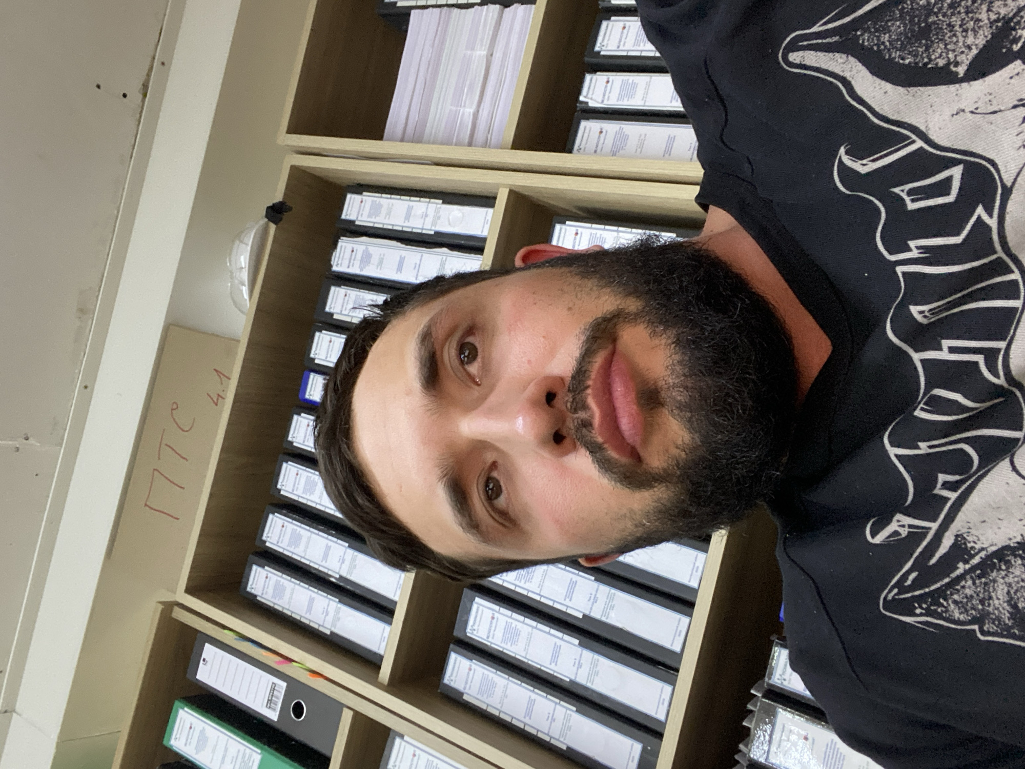 The image is a close-up, overhead shot of a man with a beard, looking upwards. He is wearing a black t-shirt with a white graphic design. Behind him, a shelf is filled with binders and stacks of paper. A sign with red Cyrillic text reading "ПТС 4.1" is visible on the shelf above the binders. The setting appears to be an office or storage space. There is no information available about the location in the city of Luzhitsy, Russia.