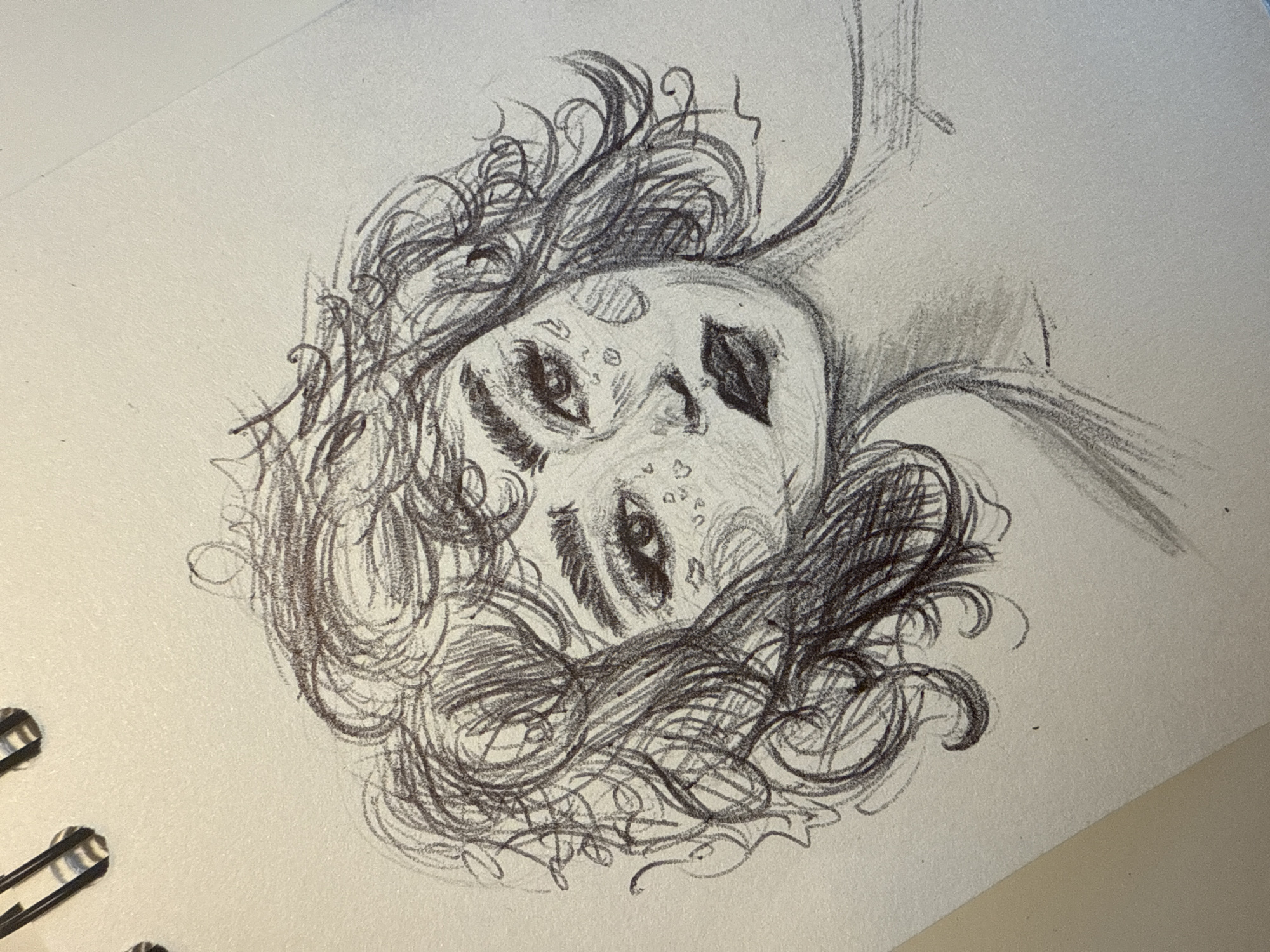 A pencil sketch on textured paper depicts a person with curly hair looking upwards. The face is stylized with exaggerated features. The left eye is detailed with eyelashes and a dark pupil, while the right eye is similar but slightly less defined. Below the right eye are small shapes resembling dots or teardrops. The nose is shaped with shading and a dark patch is drawn to the right of the nose. The hair is rendered as a mass of curls, with some strands extending outwards. The background is the white paper of the sketchbook. The location is unknown, and there is no evidence to suggest that the image was taken in Sofia, Bulgaria.