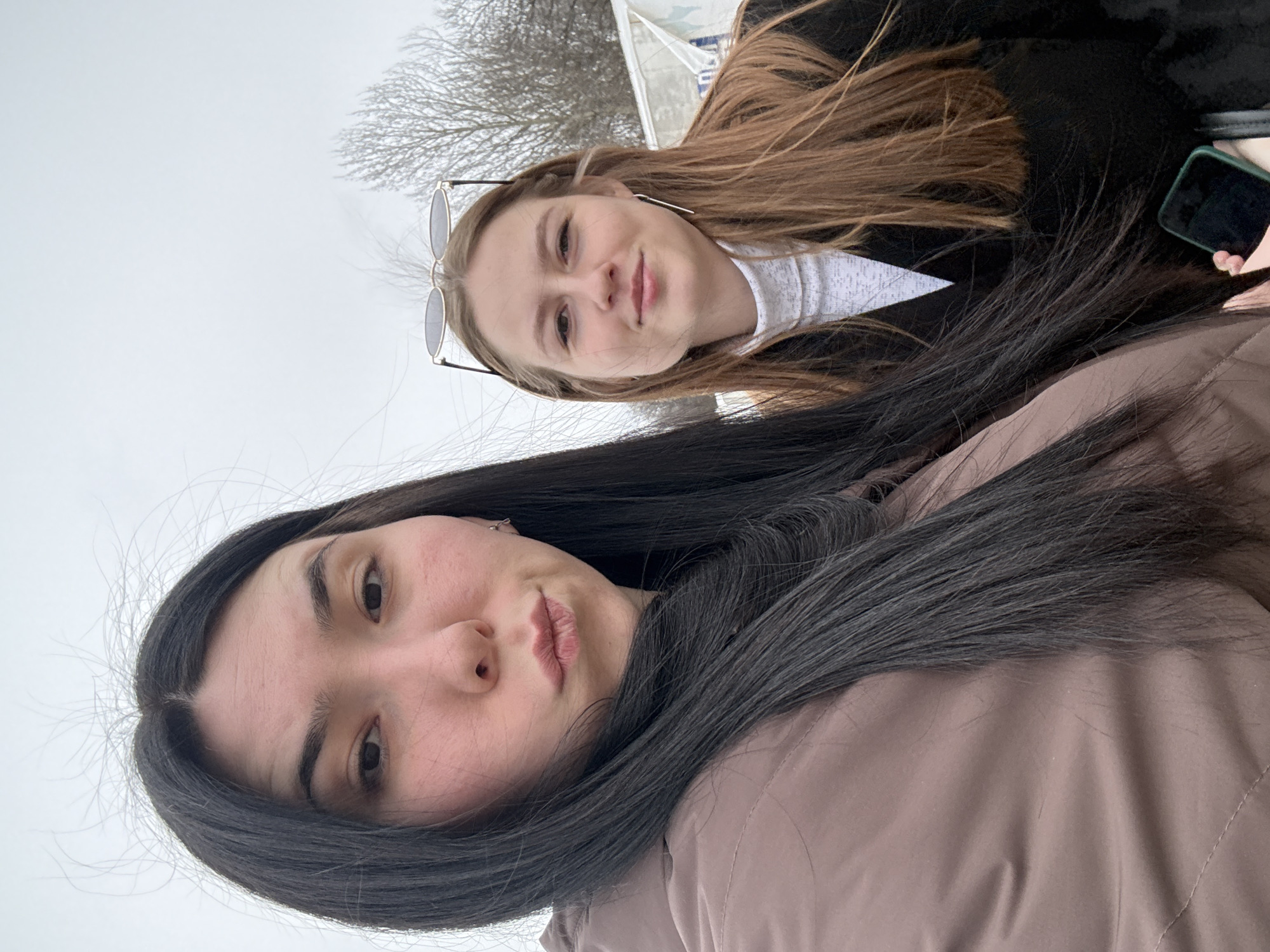 This is an outdoor selfie featuring two young women in Nizhnie Yaki, Russia, likely taken during late autumn or winter given the bare tree branches in the background. The image is oriented vertically, with the subjects appearing from a slightly downward-looking perspective.
In the foreground, a woman with long, dark, straight hair, parted down the middle, looks directly at the camera. She has a neutral expression with her lips slightly pursed and dark eyes visible. She is wearing a light brown or dusty rose-colored puffer jacket.
Behind her, partially obscured, is another young woman with long, light brown hair. She is smiling gently and also looking towards the camera. She has a pair of sunglasses pushed up onto her head and is wearing a black jacket over a white, high-necked top. Her right hand, holding a mobile phone with a dark green case, is visible on the right side of the frame.
The background consists of a bright, diffused sky, indicating an overcast day. Sparse, bare tree branches are visible against the light background on the upper left, providing a subtle environmental detail. The overall atmosphere is casual. There is no visible text within the image.