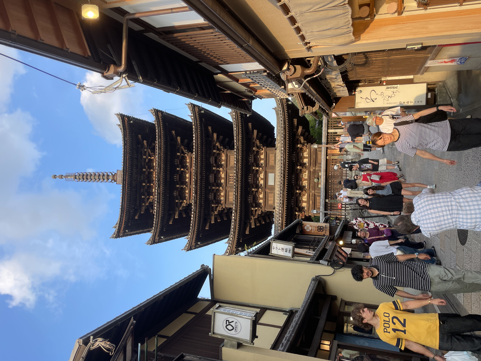 The image captures a street scene in Kyoto, Japan, with a prominent five-tiered pagoda as the focal point. The pagoda, constructed of dark wood, stands tall against a bright blue sky with scattered white clouds. The street is lined with traditional Japanese buildings with dark wooden facades and tiled roofs.
In the foreground, several people are walking along the cobblestone street. On the left, a young man wearing a yellow t-shirt with "POLO 12" printed on it is walking with his hands in his pockets. Beside him, another young man in a black and white striped polo shirt and striped pants is walking, wearing earbuds. Further down the street, a group of people are seen walking together, some dressed in casual clothing, while others are in traditional Japanese attire, such as a woman in a kimono. A man in the lower right corner is photographing the pagoda.
The street appears to be a popular tourist area, with shops and signs visible along the buildings. The lighting suggests it is late afternoon, with the sun casting long shadows and highlighting the architectural details of the pagoda and buildings. The overall atmosphere is one of a bustling yet serene historical district.