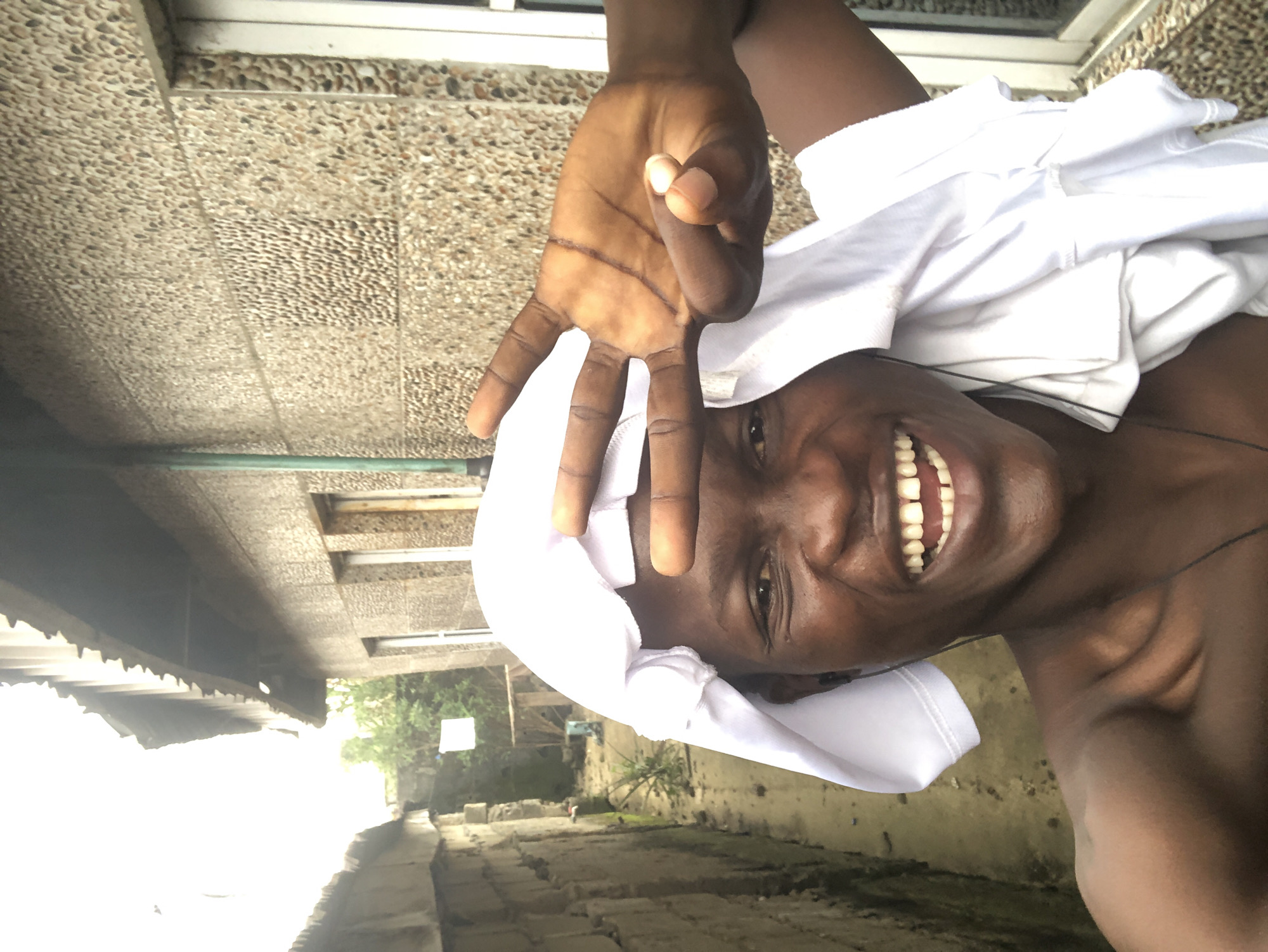 In a selfie taken in Ado, Nigeria, a young Black male with dark skin is pictured from the chest up, smiling widely. He is wearing a white cloth draped over his head and shoulders, which also partially covers his chest. His left hand is raised, with two fingers extended in a peace sign, positioned in front of his face. He appears to be wearing black earbuds. The background shows the textured, pebbled wall of a building, with a corrugated metal roof visible overhead. To the left, a narrow alleyway is partially visible, lined with a brick wall and some sparse vegetation. A window with white trim is visible on the right side of the building. The lighting suggests an overcast day.