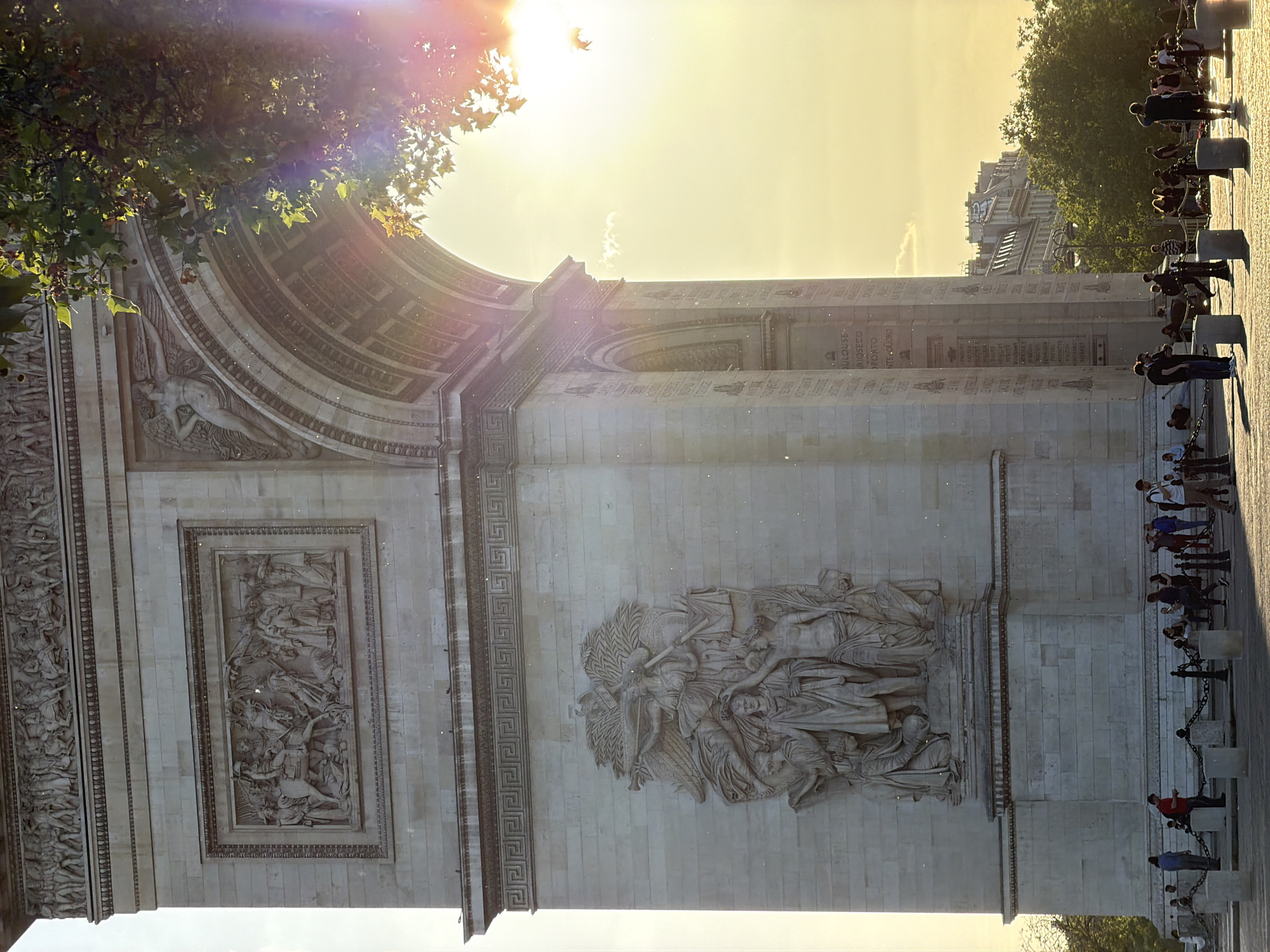 This is a daytime, outdoor, rotated photograph of the Arc de Triomphe in Paris, France. The sun is high in the sky, shining brightly, with a tree branch visible in the upper left corner. The Arc de Triomphe is made of stone and features detailed carvings on its surface. Several people are standing behind a chain fence on the right side of the image. Buildings and trees are visible in the background behind the people.