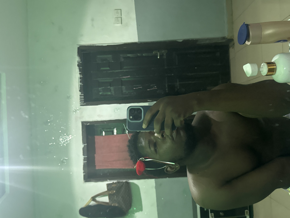 A male individual with dark skin and facial hair, shirtless, is seen taking a mirror selfie in Ipetumodu, Nigeria. He holds a dark-colored smartphone (an iPhone with a triple-lens camera system) in his right hand, partially obscuring his face. A red ornamental headband is worn on his head. On a surface directly beneath the mirror, two bottles are present: one white with a gold pump dispenser and a white cap, and another brown with a blue flip-top cap. The background, reflected in the visibly speckled mirror, features light green walls. Two dark wooden doors are visible; the left door is partially ajar, revealing a red curtain behind it. A brown, patterned handbag hangs on the wall to the left, and two white light switches are mounted on the wall to the right. This scene is located within a private residence in Ipetumodu, Nigeria.