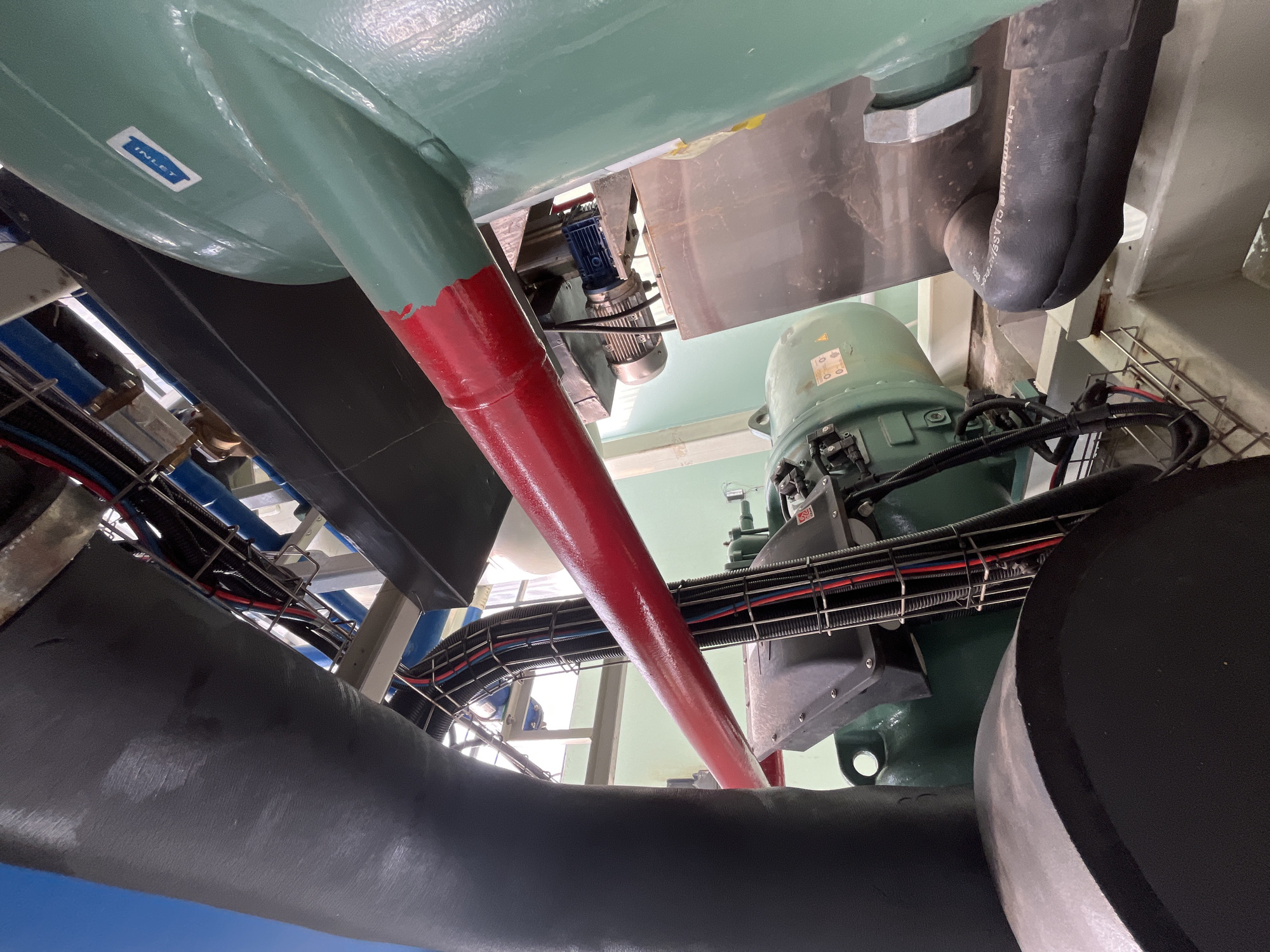 The image displays a close-up view of industrial machinery, likely part of a cooling or HVAC system, situated in Pekalongan, Indonesia. Prominently featured are large, insulated pipes, one painted red and another green. A dark, flexible hose with the text "HUMAK KG CLASS E2" is visible in the lower right, connected to a green cylindrical component. A green, industrial motor with black wiring protected by a ribbed conduit is mounted below a section of the red pipe. The background shows more piping, electrical conduits, and structural elements of an industrial setting. No people are visible in the frame. The lighting suggests an indoor or covered industrial space.