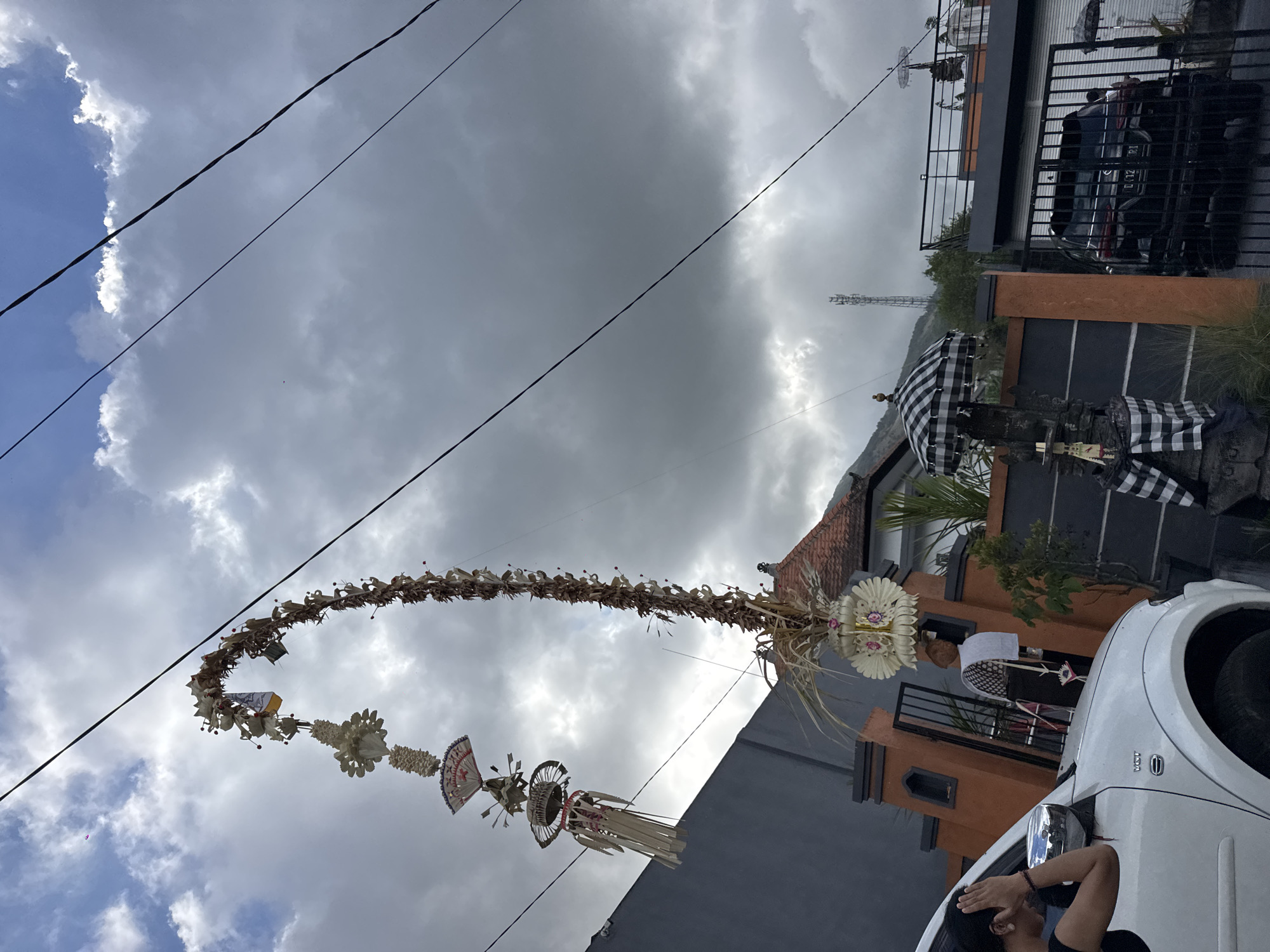 The image shows an outdoor scene in Kintamani, Indonesia, under a cloudy sky with some patches of blue. A prominent feature is a tall, arched decorative structure made of natural materials, possibly bamboo and dried leaves, adorned with white floral-like ornaments and flags. This structure appears to be suspended from overhead wires, which are a noticeable element in the sky.
In the foreground, a white SUV is parked. A person, appearing to be a woman with dark hair, is partially visible inside the SUV on the driver's side, with her hand near her face, looking towards the left.
Behind the car, there is a building with an orange-tiled roof and a dark-colored wall. An entrance gate to this property is visible, with a black metal gate. To the right of the gate, a black SUV is parked behind the gate.
Further to the right, there are more buildings and a prominent black and white checkered umbrella, typical of Balinese religious ceremonies, placed on a stone structure. Another similar umbrella is visible on a balcony on the upper floor of a building. A cellular tower can be seen in the background on a hill. The overall impression is a street scene with traditional Balinese decorations.