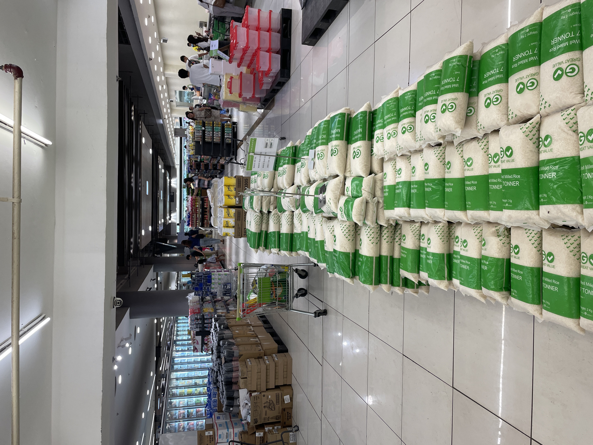 This image depicts the interior of a supermarket, likely located in Davao City, Philippines, based on the "Gaigano Market" sign visible on a price tag. The scene is dominated by rows of stacked bags of rice, predominantly white with green packaging, arranged in long lines on the tiled floor. Several shopping carts are present, some empty and some containing items, indicating customer activity. In the background, shelves stocked with various products, including beverages in coolers and other packaged goods, are visible. A few individuals, including shoppers and possibly store employees, are scattered throughout the store, some pushing carts or browsing shelves. The overall environment is a typical retail setting with fluorescent lighting overhead.