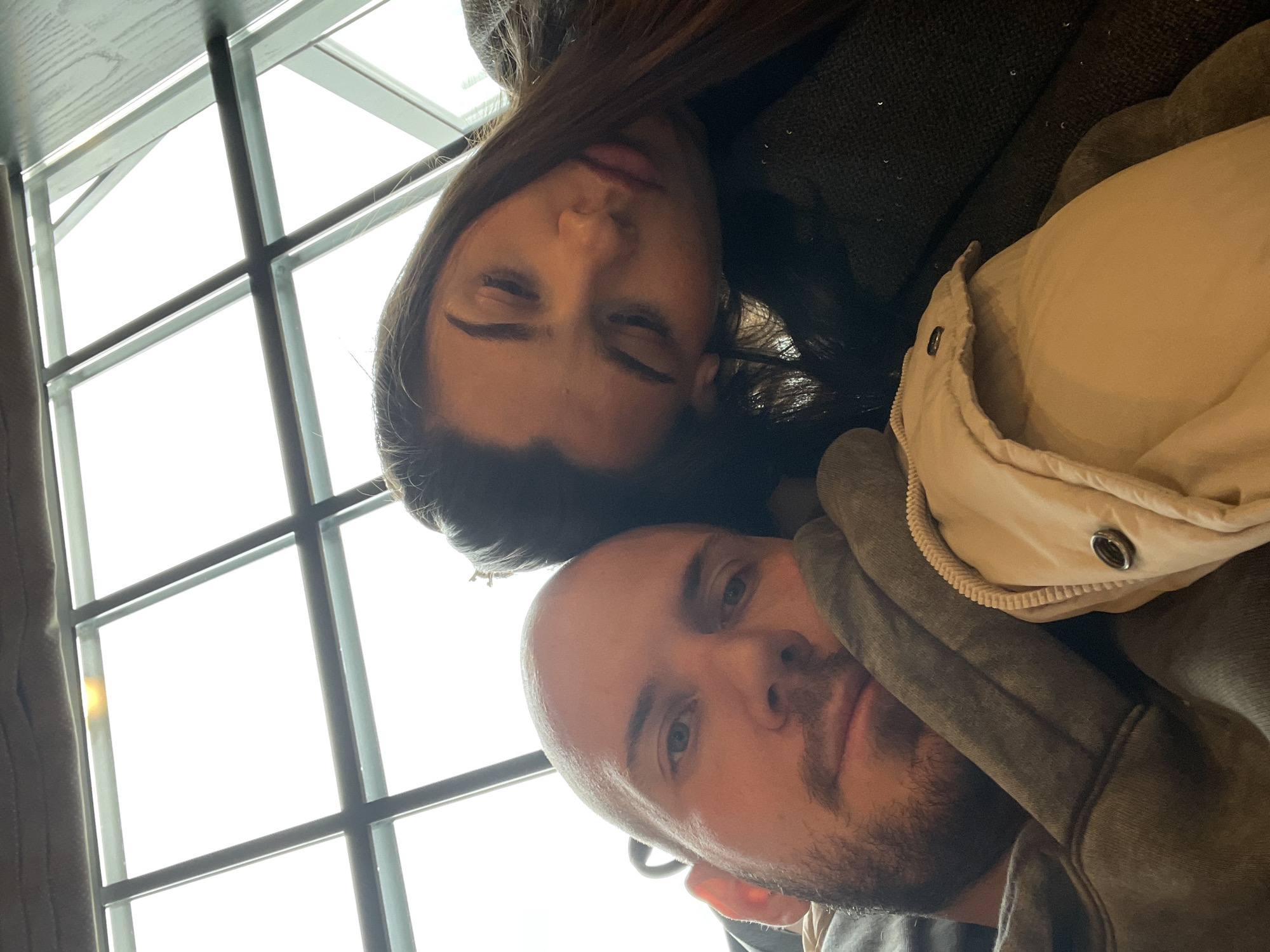The image shows two individuals, a woman and a man, posing together. The woman, with long, dark hair, has her eyes closed. The man is bald with a beard and is looking at the camera. The background is a window with a view to the outside. The location appears to be inside, and it can not be determined that it is in Krasnoyarsk, Russia.