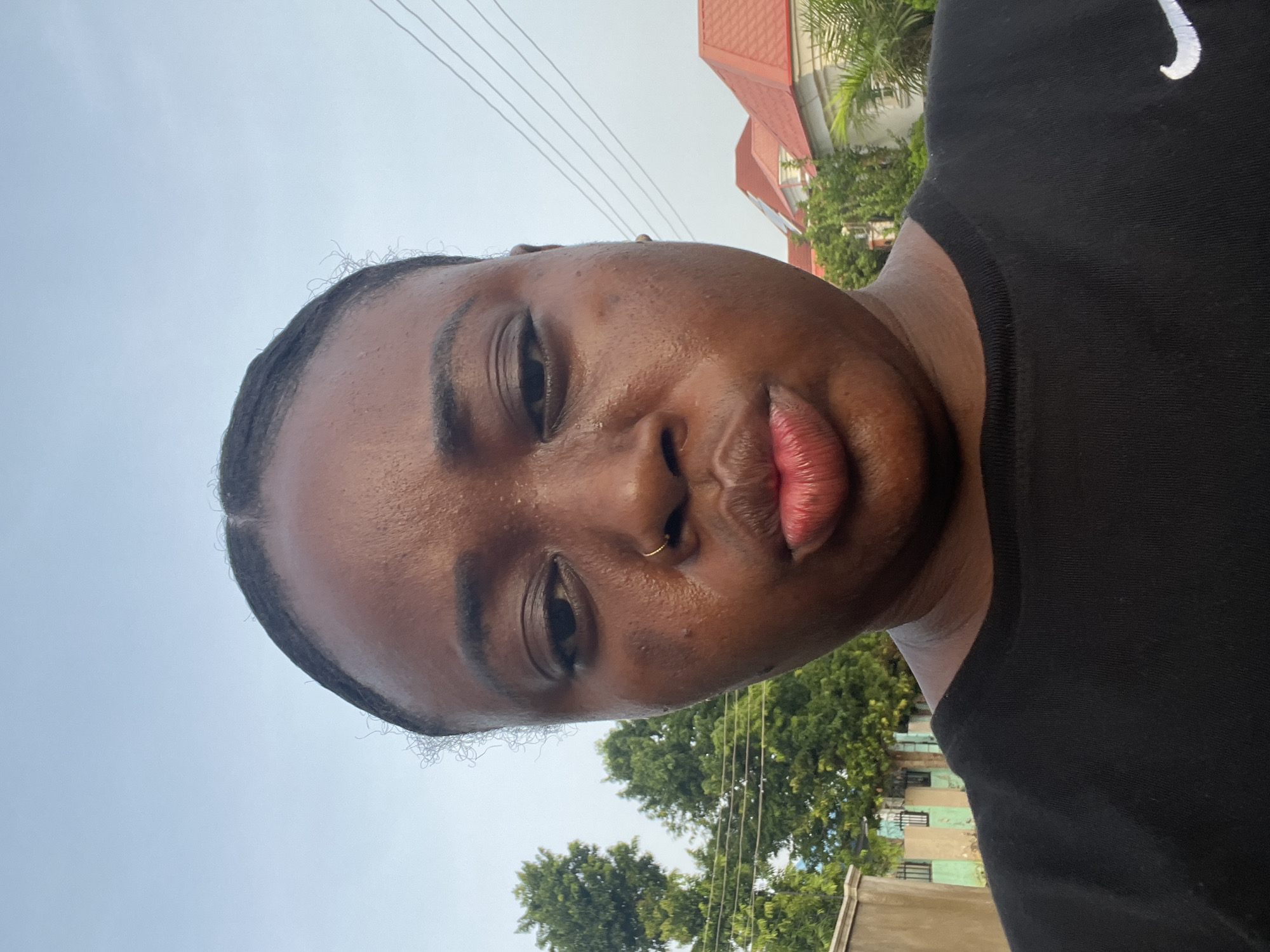 A selfie taken outdoors shows a dark-skinned person with a nose ring, wearing a black Nike t-shirt. Buildings with red roofs and green trees are in the background. The location is potentially in Abuja, Nigeria, given the prompt context. Overhead power lines are visible. The person appears to be in the sunlight.