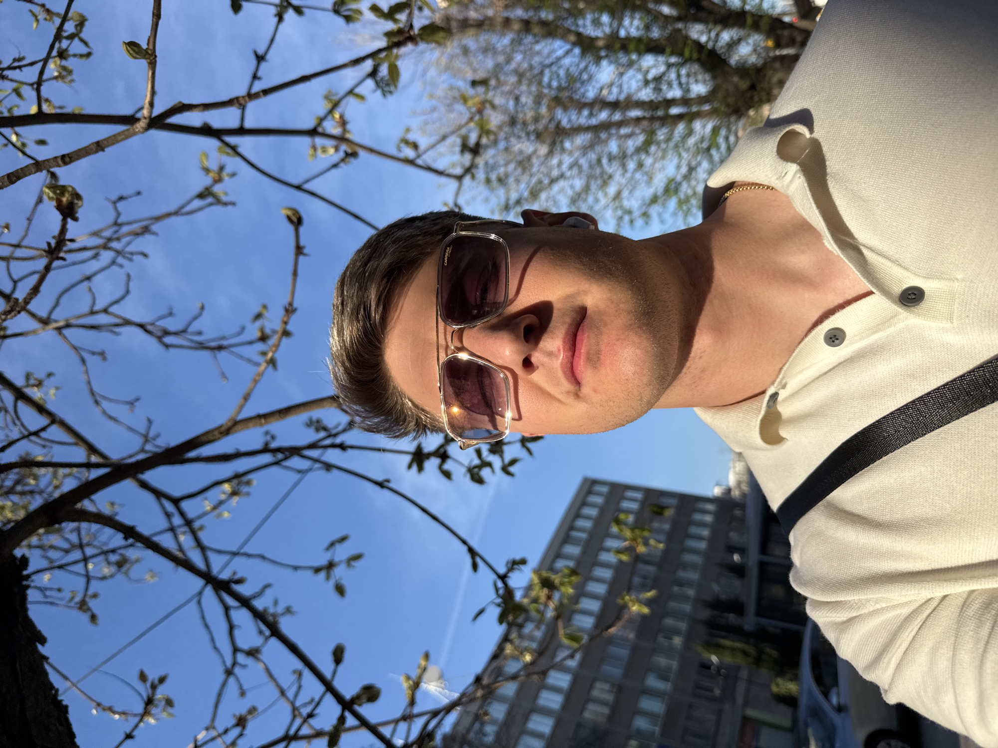 A medium shot shows a man with short brown hair and sunglasses, facing downward towards the camera. He is wearing a white polo shirt with a black strap running across his torso. A gold chain is visible around his neck. Behind him and slightly to the left are branches of a tree with budding leaves. The background includes a clear blue sky, a multi-story building with windows, and a blurred car. The overall impression suggests an outdoor location in an urban environment. There is no information to determine that the location is in Plovdiv, Bulgaria.
