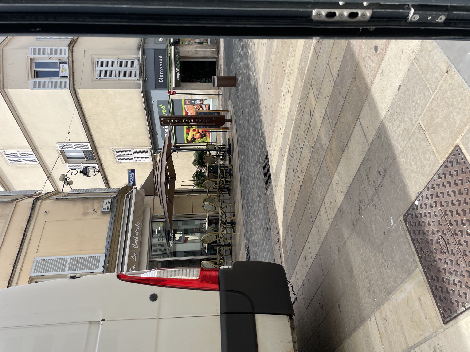 The image shows a street scene in Meaux, France. In the foreground, partially visible on the left, is the rear of a white van. The street is paved with light-colored stone tiles, with a darker cobblestone area visible further back. On the right side of the image, a dark metal frame is present.
In the mid-ground, there is an outdoor seating area for a brasserie, with several round tables and chairs set up under a large umbrella. The brasserie is identified by a sign reading "De La Cathédrale". Adjacent to the brasserie is a "Rapid Market" store with a sign that says "Bienvenue".
In the background, buildings line the street, featuring typical French architecture with shutters on the windows. A street sign labeled "RUE SAINT REMY" is visible. The scene appears to be a pleasant, sunny day.