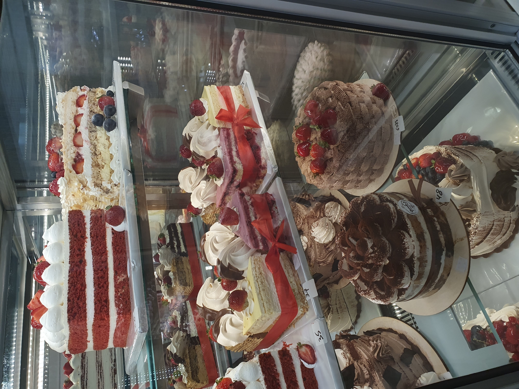 A display case filled with various cakes is shown in this medium-shot, eye-level photograph. The cakes are arranged on multiple shelves within a refrigerated glass display case, suggesting a bakery or pastry shop.
The top shelf features a tall, square red velvet cake topped with whipped cream and strawberries, and a layered cake with strawberries, blueberries, and whipped cream.
The middle shelf showcases several slices of cake and smaller whole cakes. One prominent item is a slice of cake layered with yellow cream and red velvet, adorned with a red ribbon and fresh strawberries. Other cakes on this shelf include a slice of chocolate and cream cake, and a slice of pink cake with cream and strawberries.
The bottom shelf displays several whole cakes. A prominent cake is a round chocolate cake decorated with a basketweave pattern of chocolate frosting and topped with fresh strawberries. To its left, a tiramisu-style cake is covered in whipped cream and cocoa powder. Another cake to the left of the tiramisu appears to be a layered chocolate cake. In the foreground, partially visible, are more cakes with cream and fruit toppings, as well as some smaller meringue-like pastries.
The overall impression is one of a well-stocked confectionery, highlighting a variety of sweet treats. The image does not contain any people or explicit location markers within the frame itself, but if this display is located in Batumi, Georgia, it is likely in a bakery such as "Seran Bakery" or a similar establishment that sells such desserts. The lighting is bright and even, typical of a retail display case.