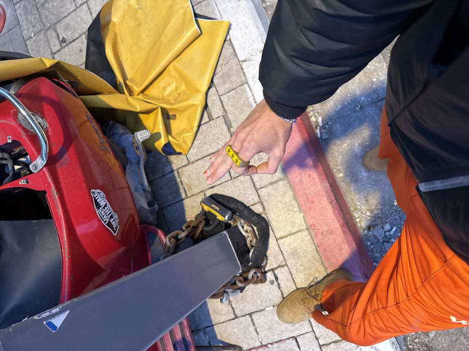 A person stands on a brick sidewalk next to a red scooter. They wear orange pants and tan boots. On their right hand, they have a gold ring with three stars on it. The scooter is locked with a black and yellow lock, as well as a chain. Next to the scooter is a large piece of yellow fabric. The person has a black jacket on.